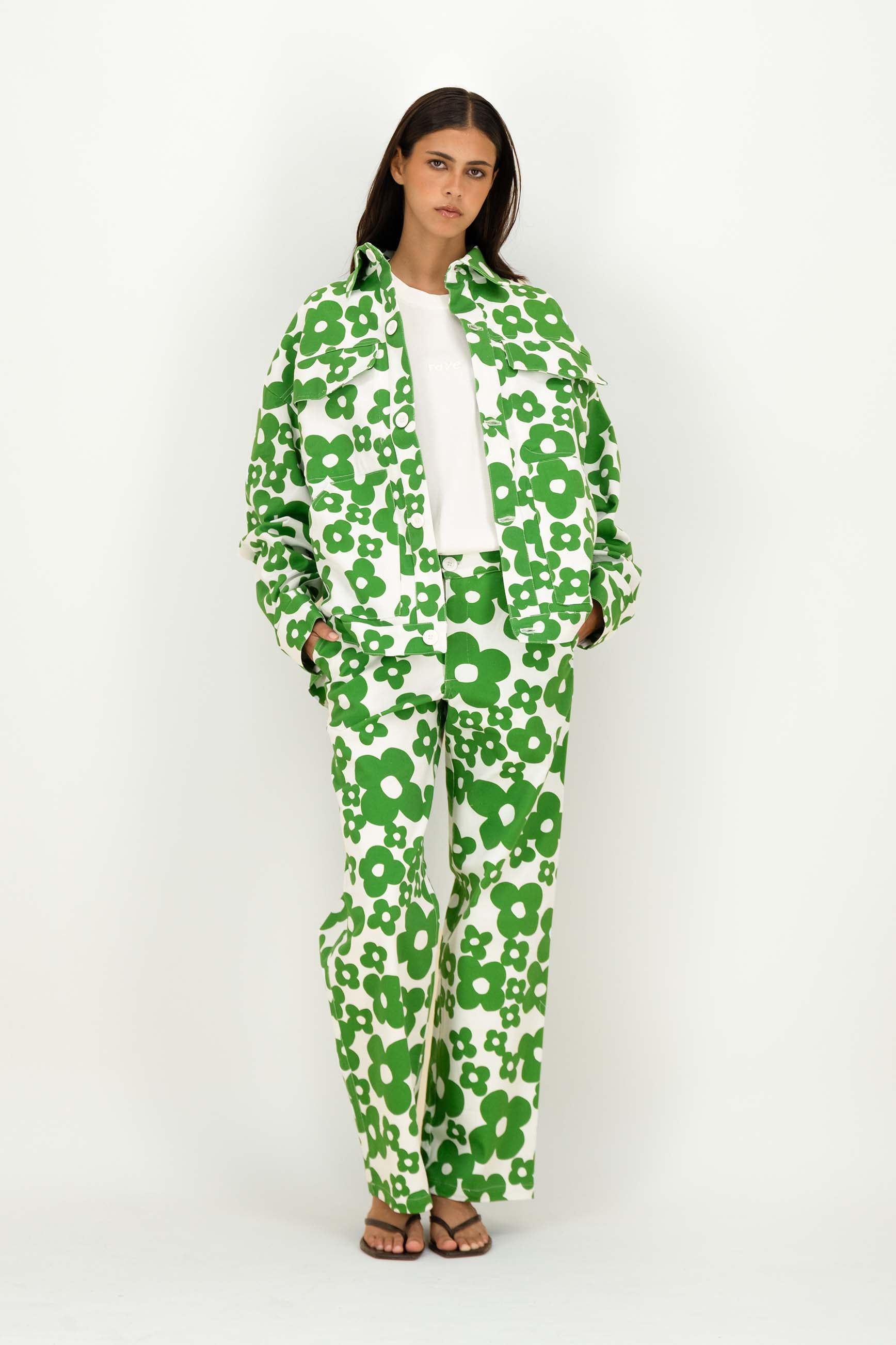 A woman modeling a matching green and white floral print jacket and trousers set, worn over a white top and brown sandals, against a plain white studio background.