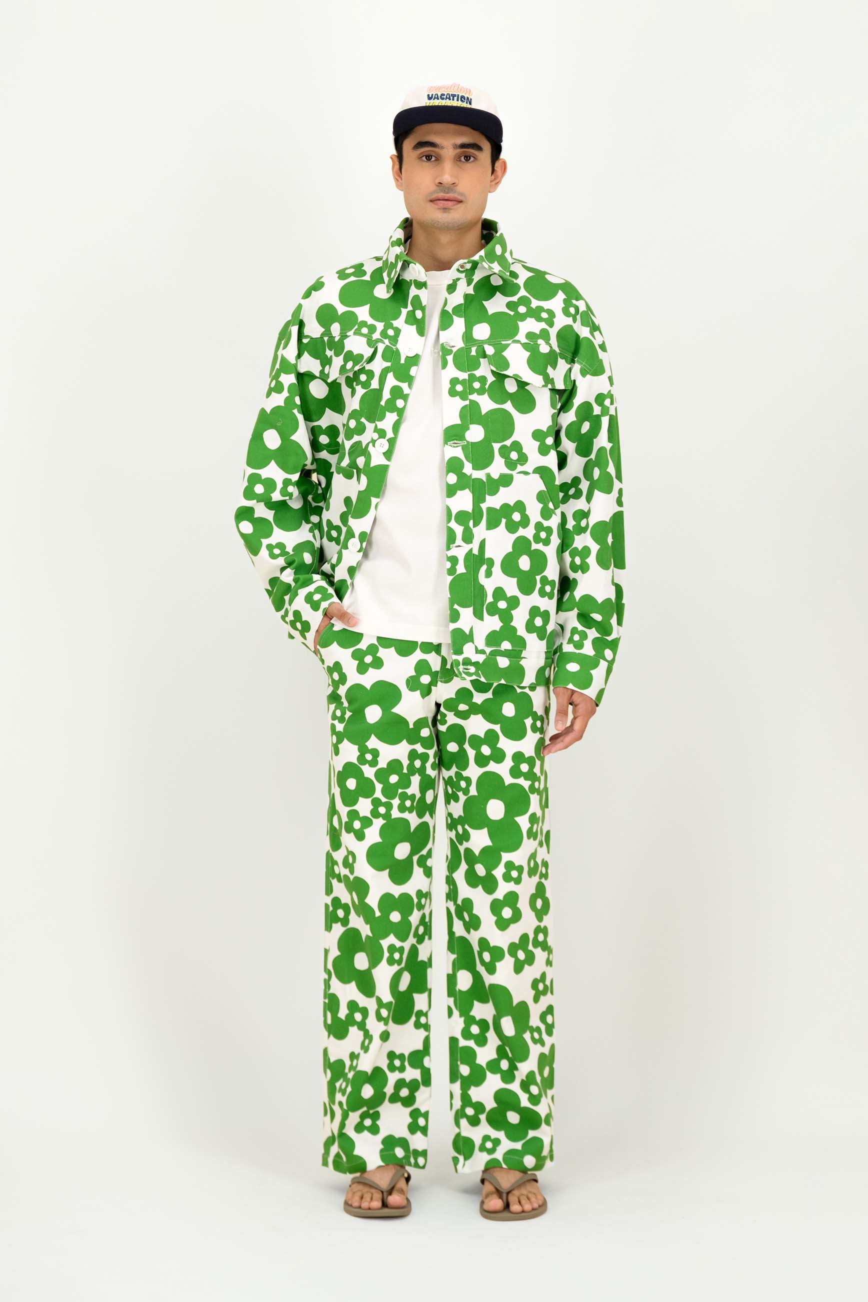 A man modeling a matching green and white floral print set, styled with a white t-shirt, brown sandals, and a black baseball cap.