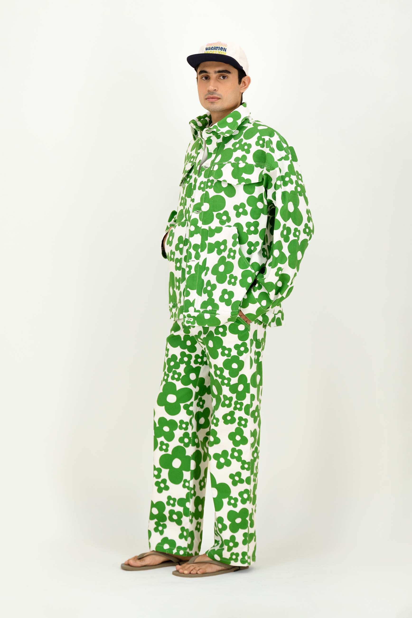 A man modeling a matching green and white floral print set and wide-leg pants, paired with a black and white baseball cap and brown sandals, against a plain white studio background.