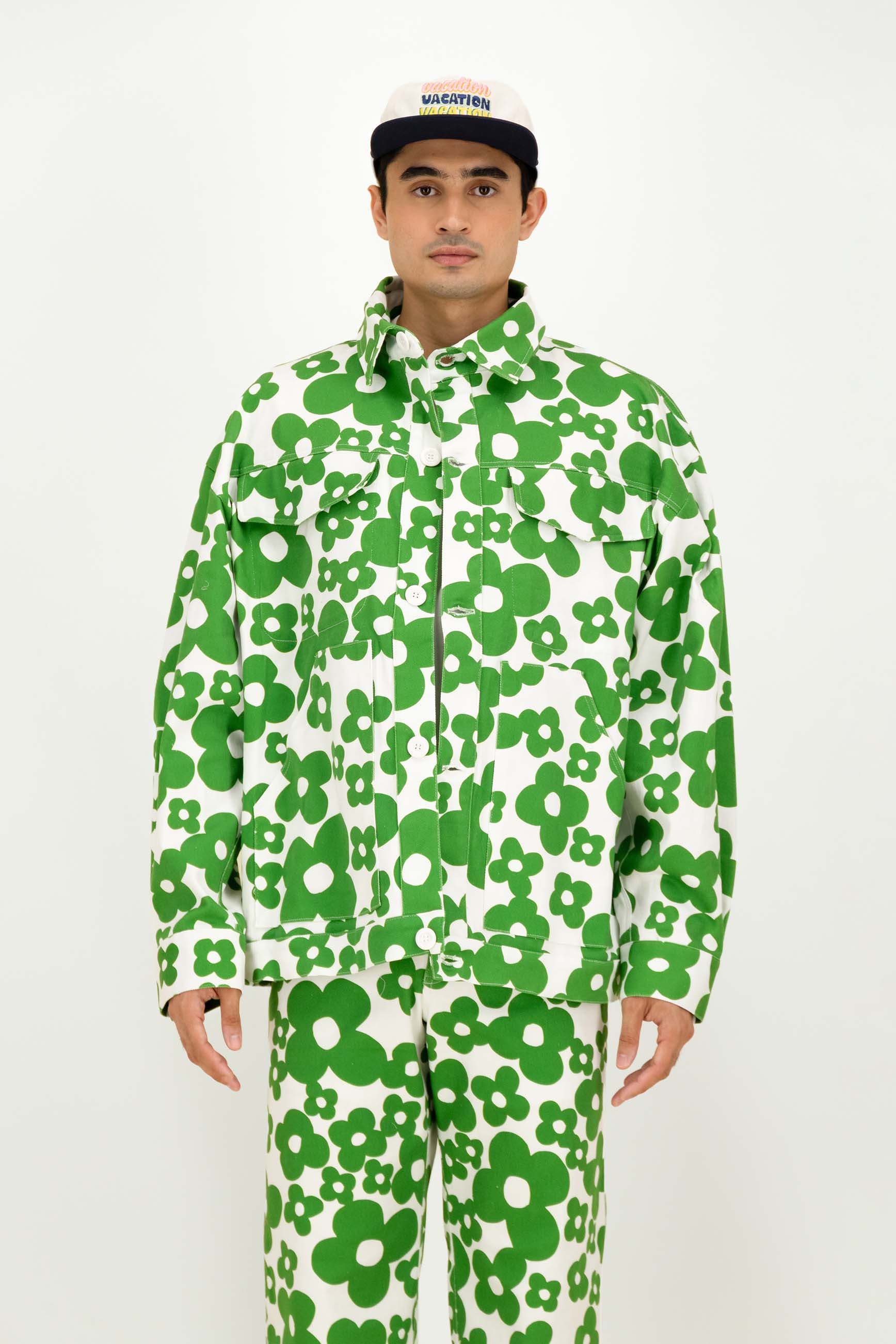 A man modeling a matching green and white floral print jacket and pants set, paired with a black and white cap, against a plain white studio background.