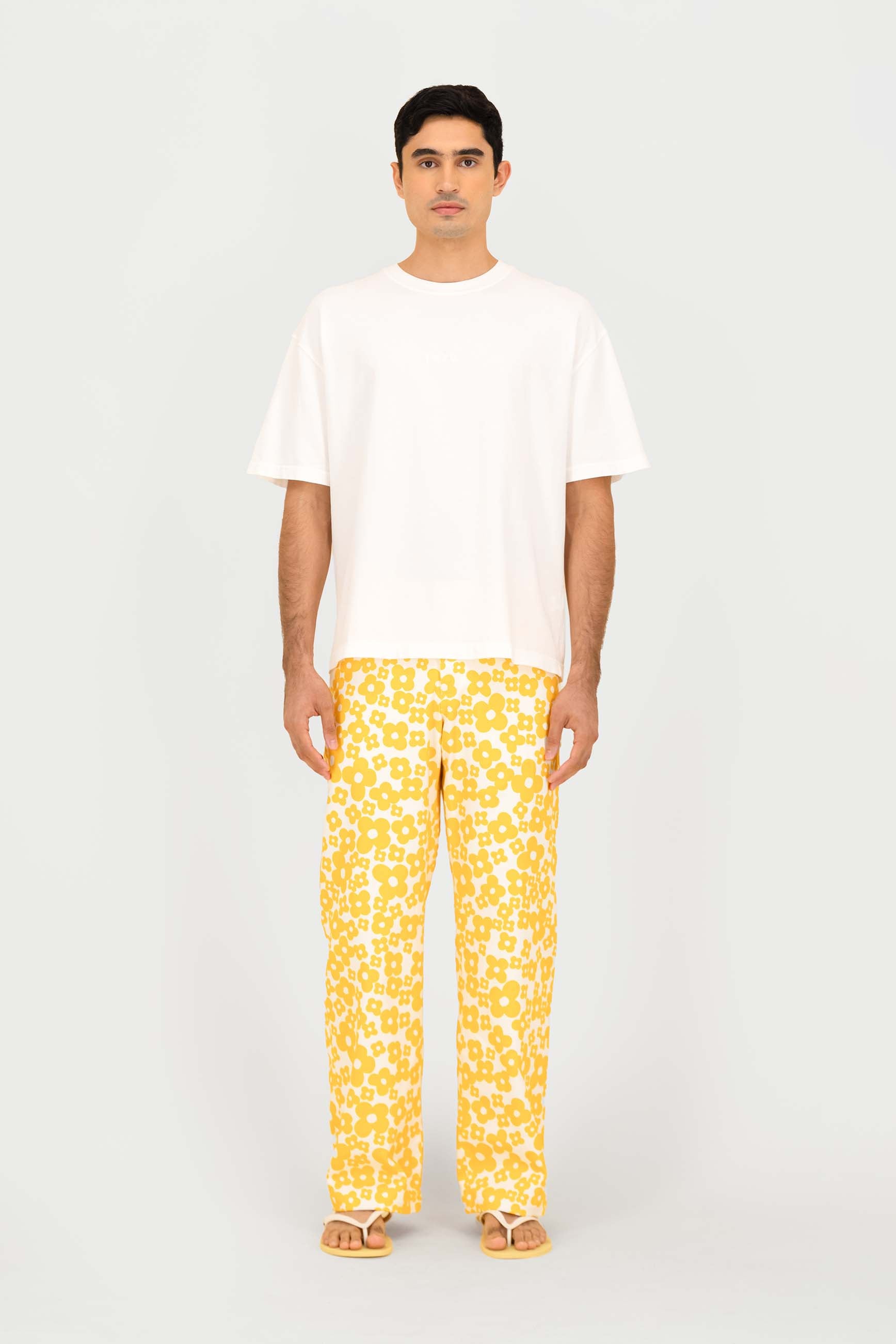 A man stands against a light gray studio background wearing a plain white short-sleeve T-shirt, bright yellow patterned pants, and tan sandals.
