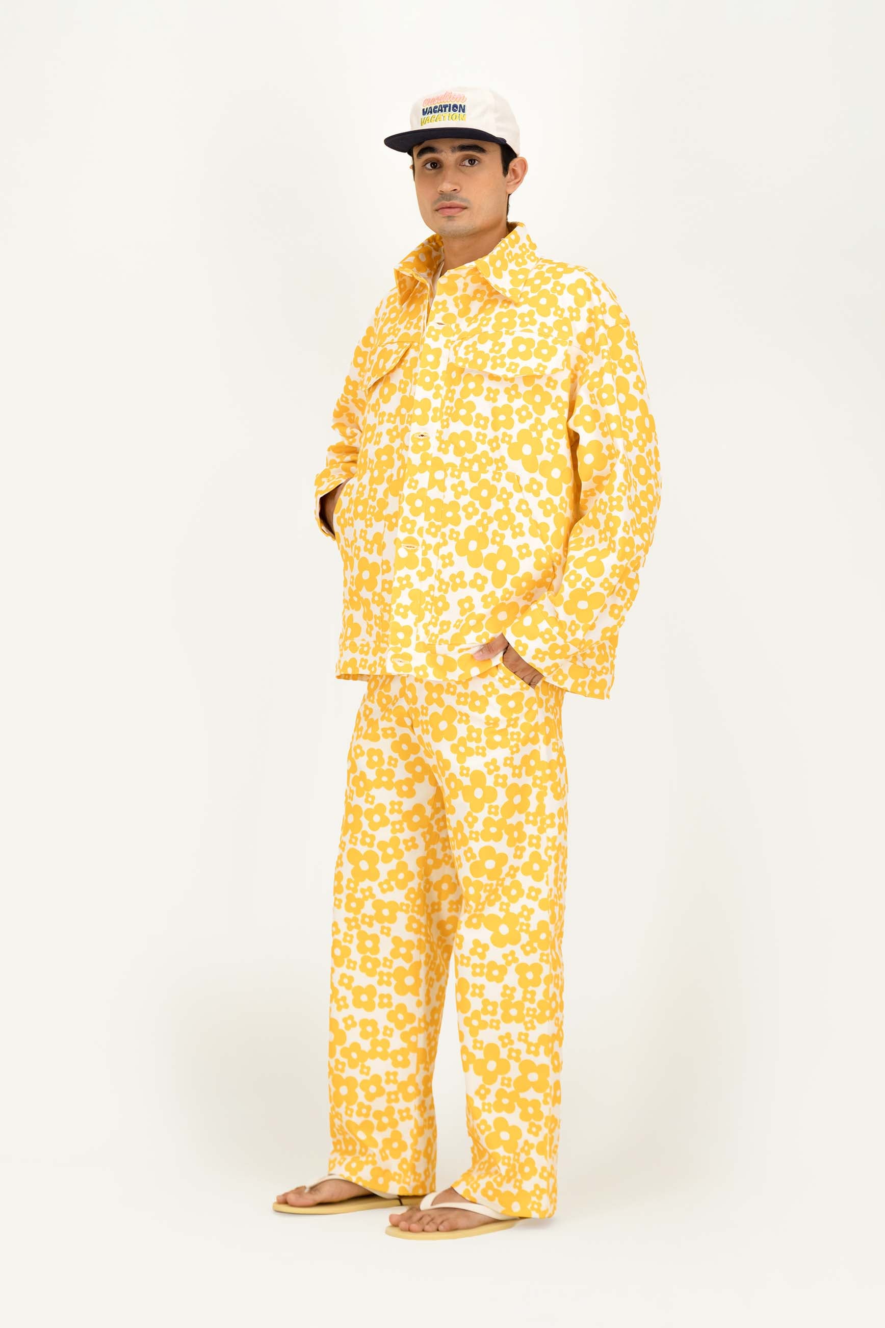 A man modeling a matching two-piece pajama set in a bright yellow and white floral pattern, paired with a white baseball cap and tan sandals, against a plain white studio background.