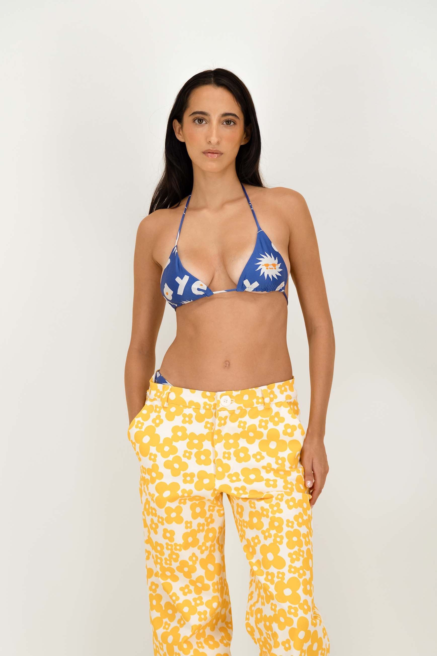 A woman modeling a two-piece swimsuit set consisting of a blue triangle bikini top with a white sun and letter print, paired with yellow and white floral print pants, against a plain light background.