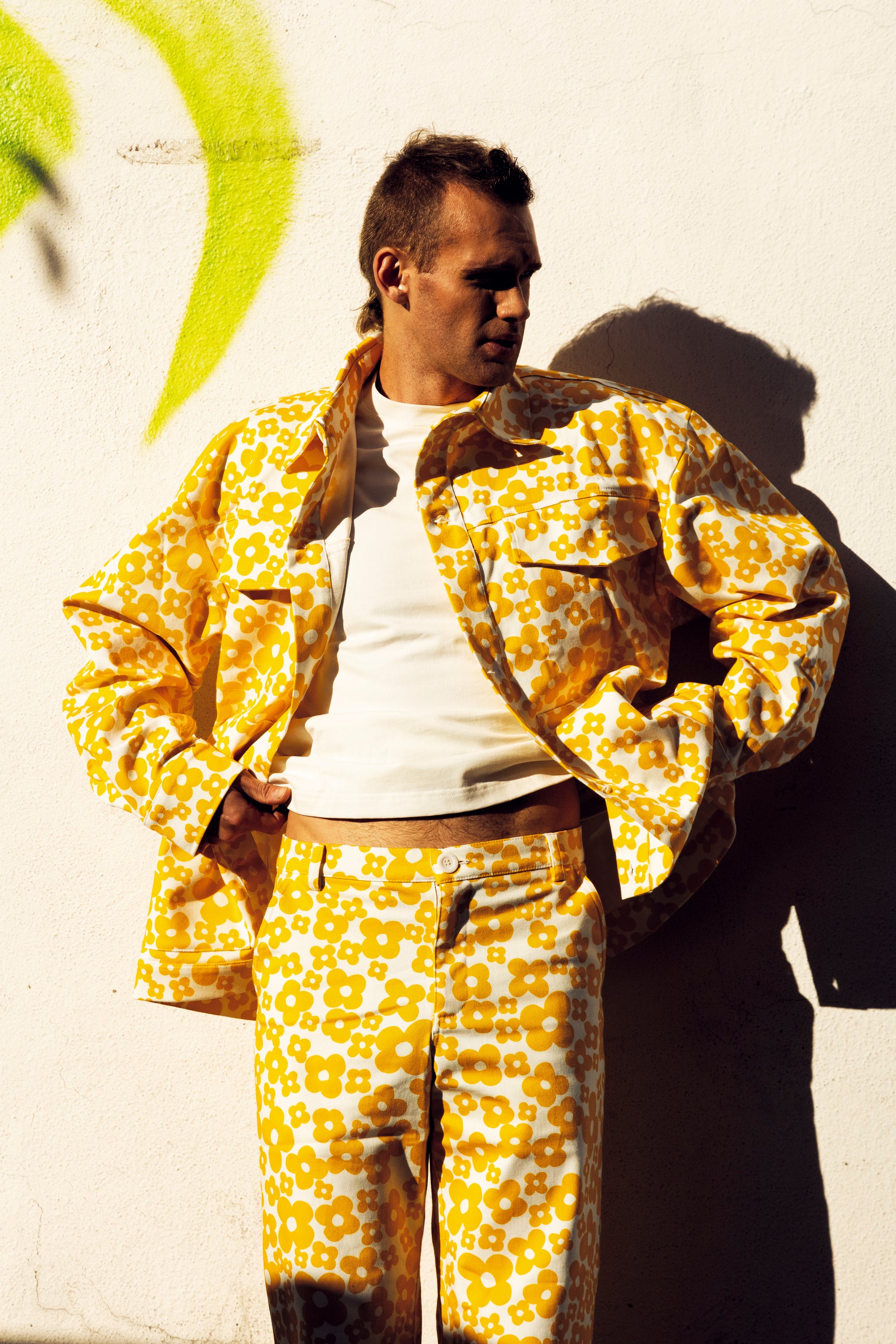 A man wearing a matching yellow and white floral print jacket and trousers set, styled over a white t-shirt, standing against a white wall with a strong shadow.