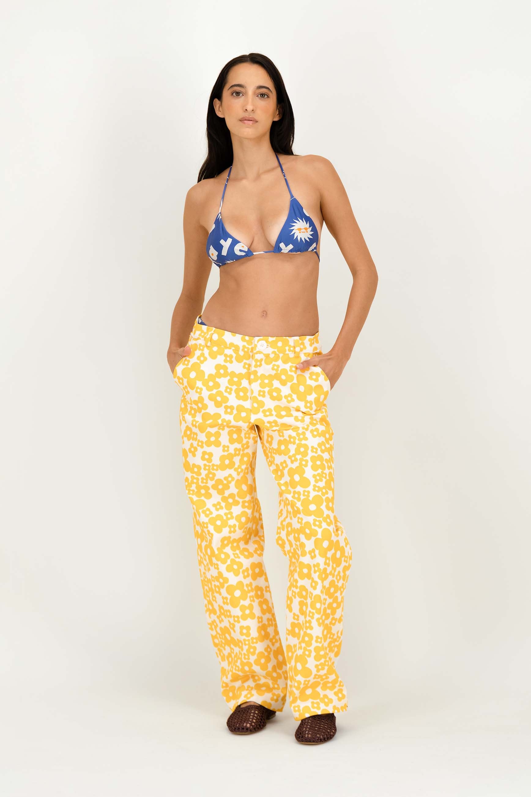 A woman modeling a two-piece swimsuit with a blue and white floral triangle bikini top and yellow-and-white patterned wide-leg pants, standing against a plain light background.