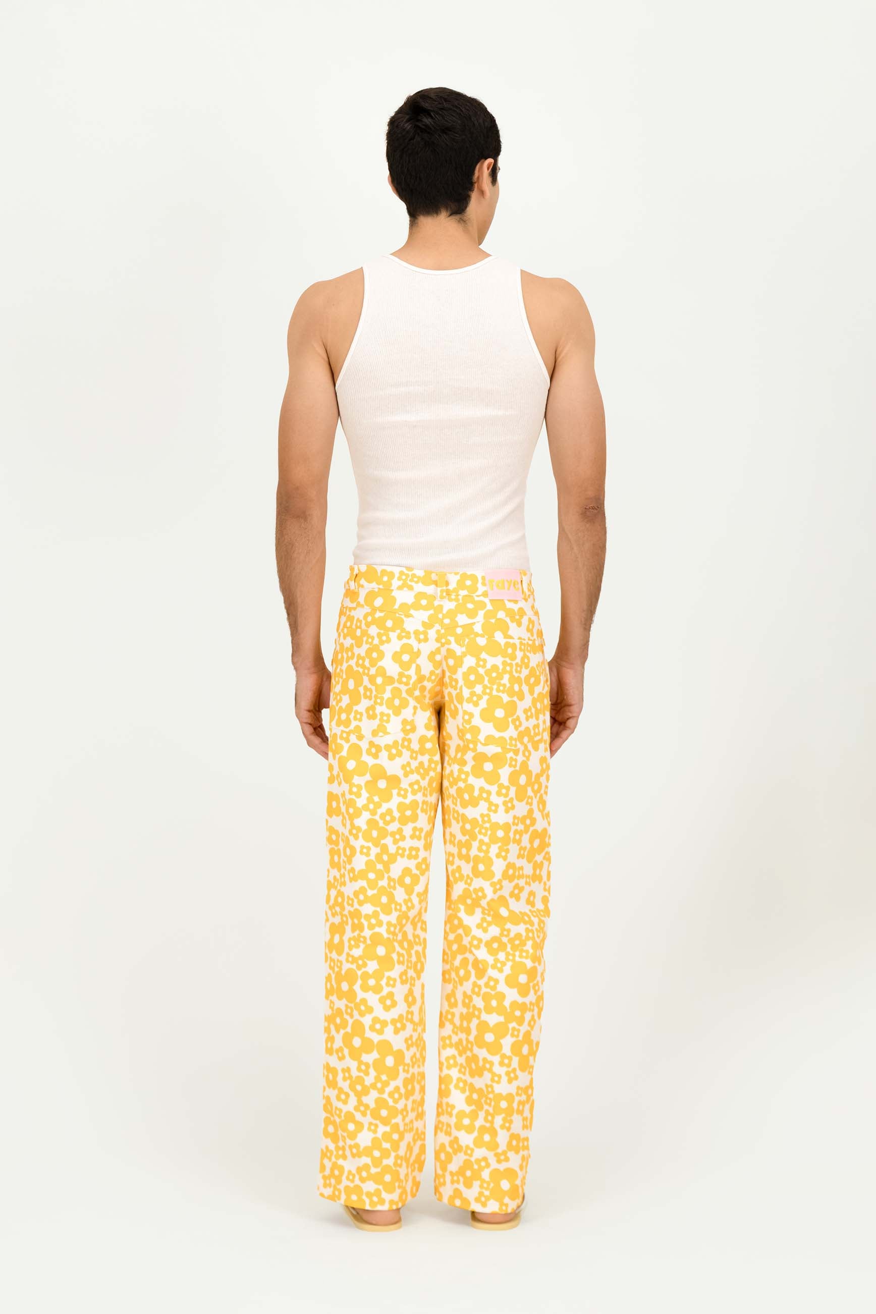 A man viewed from behind wearing a white tank top and yellow wide-leg trousers with a white abstract pattern, standing against a plain white studio background.