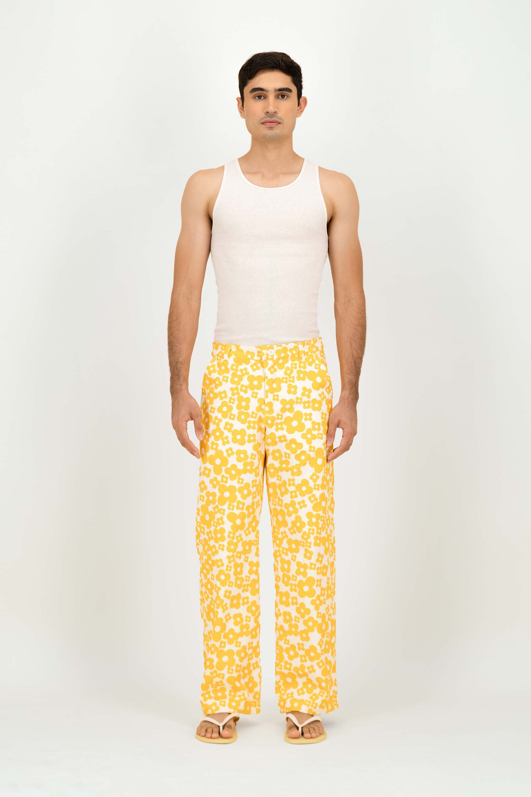 A man modeling a pair of yellow and white patterned trousers with a white tank top, standing against a plain white studio background.