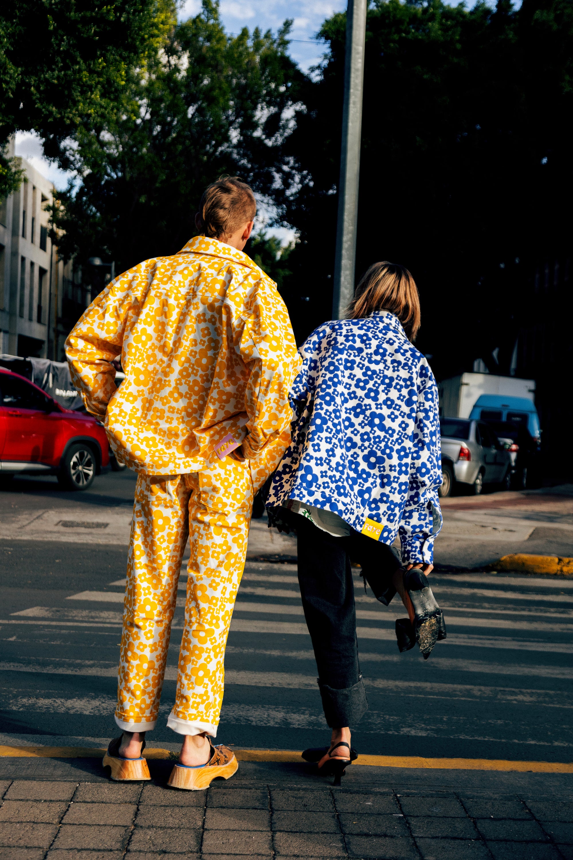 Two people seen from behind walking across a street crosswalk. The person on the left wears a bright yellow and white floral patterned jacket and matching pants, while the person on the right wears a blue and white floral patterned jacket, dark pants, and sandals.