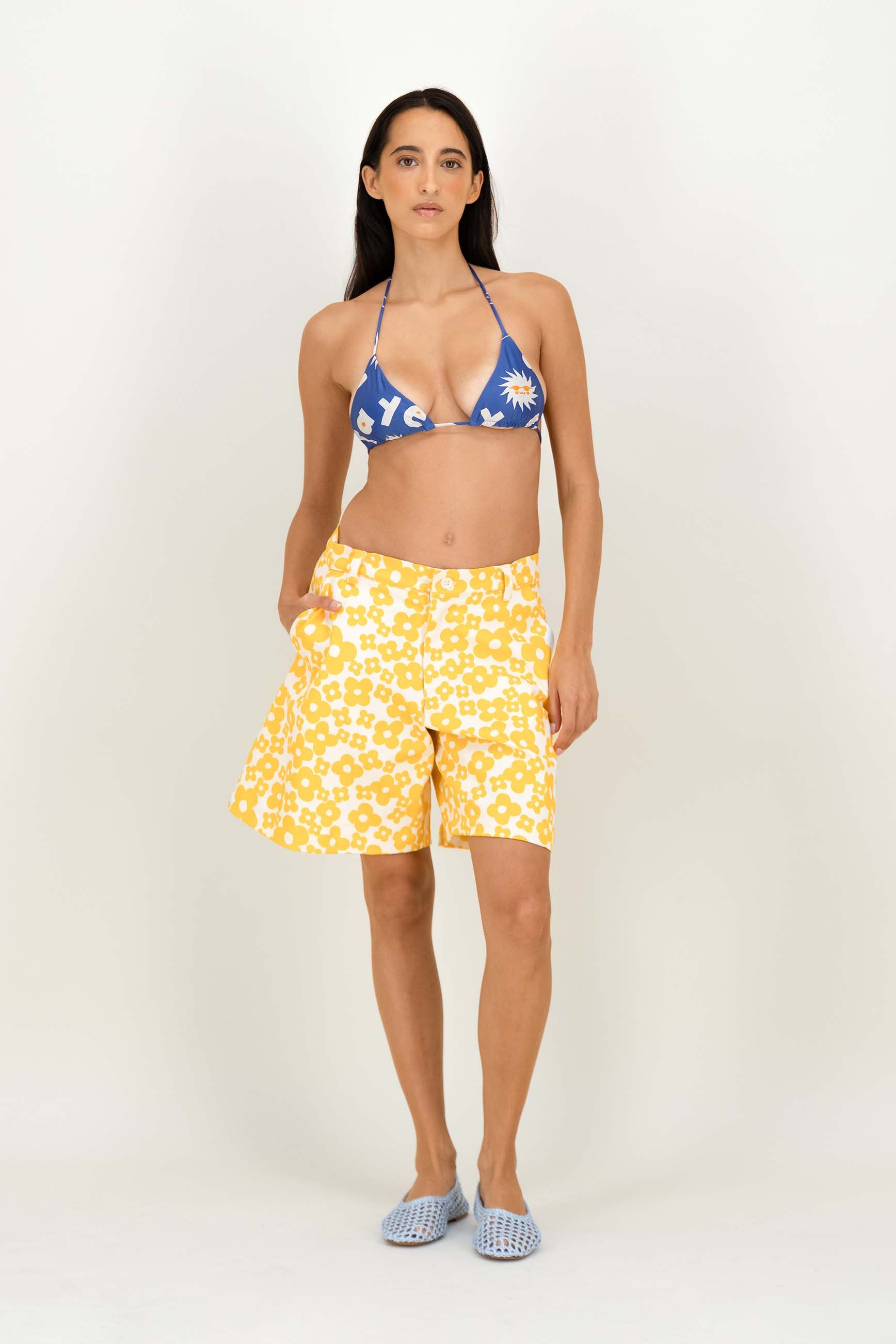 A woman modeling a two-piece swimwear set consisting of a blue triangle bikini top with white sun and letter patterns, and yellow patterned shorts, paired with light blue slip-on shoes against a plain white studio background.