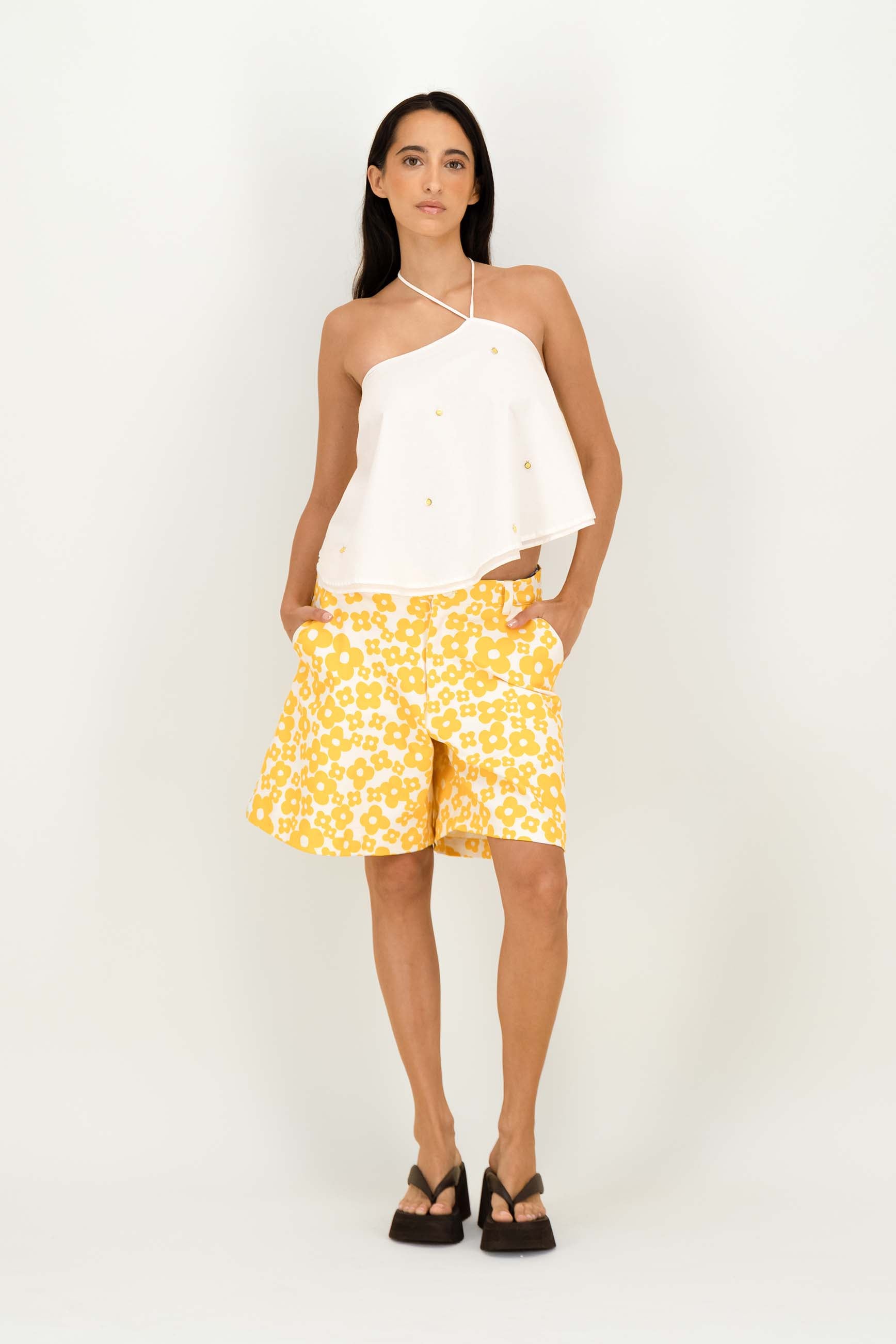 A woman modeling a white halter-neck top and yellow shorts with a white abstract pattern, standing against a plain white studio background.