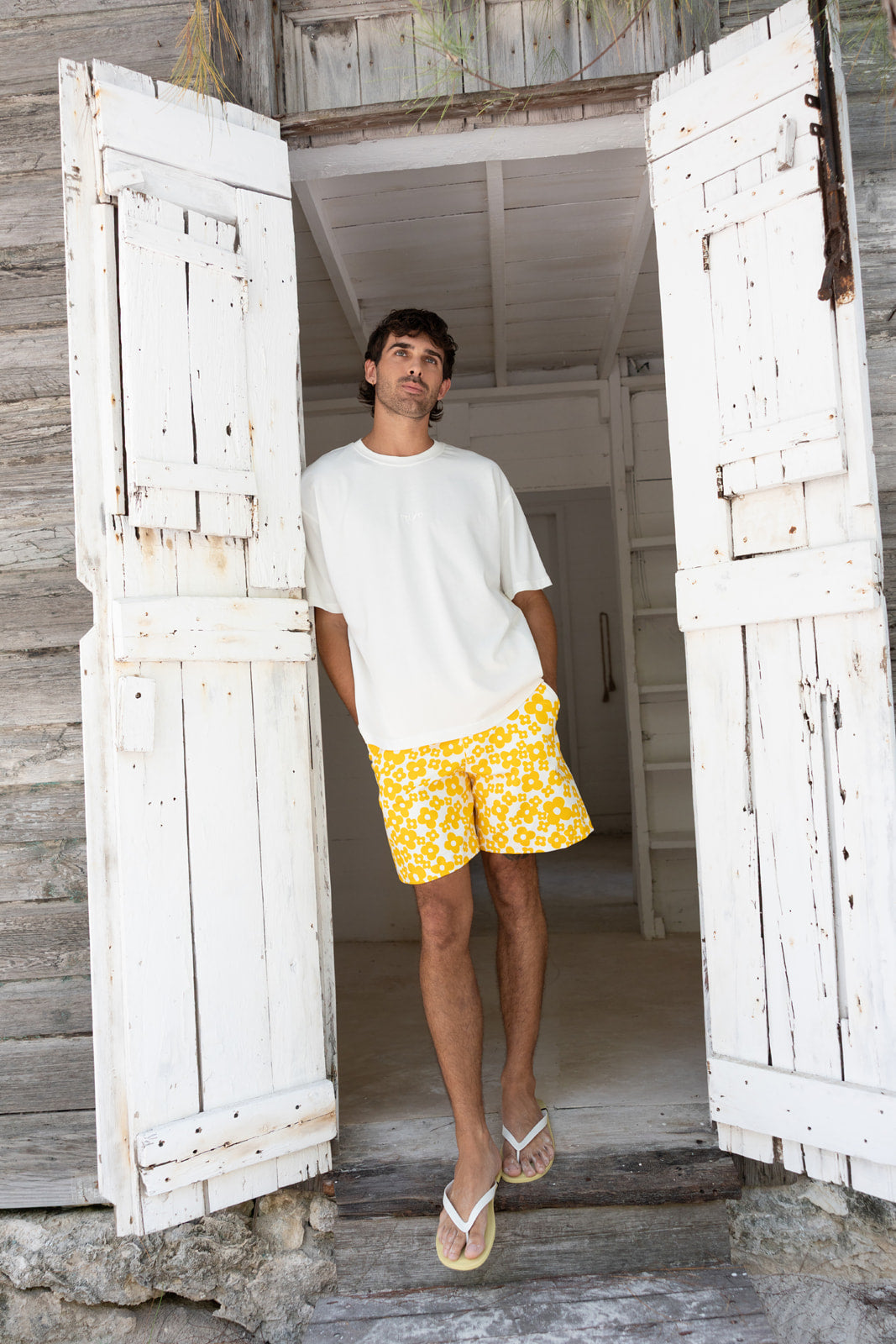 A man stands in a rustic doorway wearing a white t-shirt, yellow and white patterned swim trunks, and flip-flops, with weathered white wooden doors framing the scene.