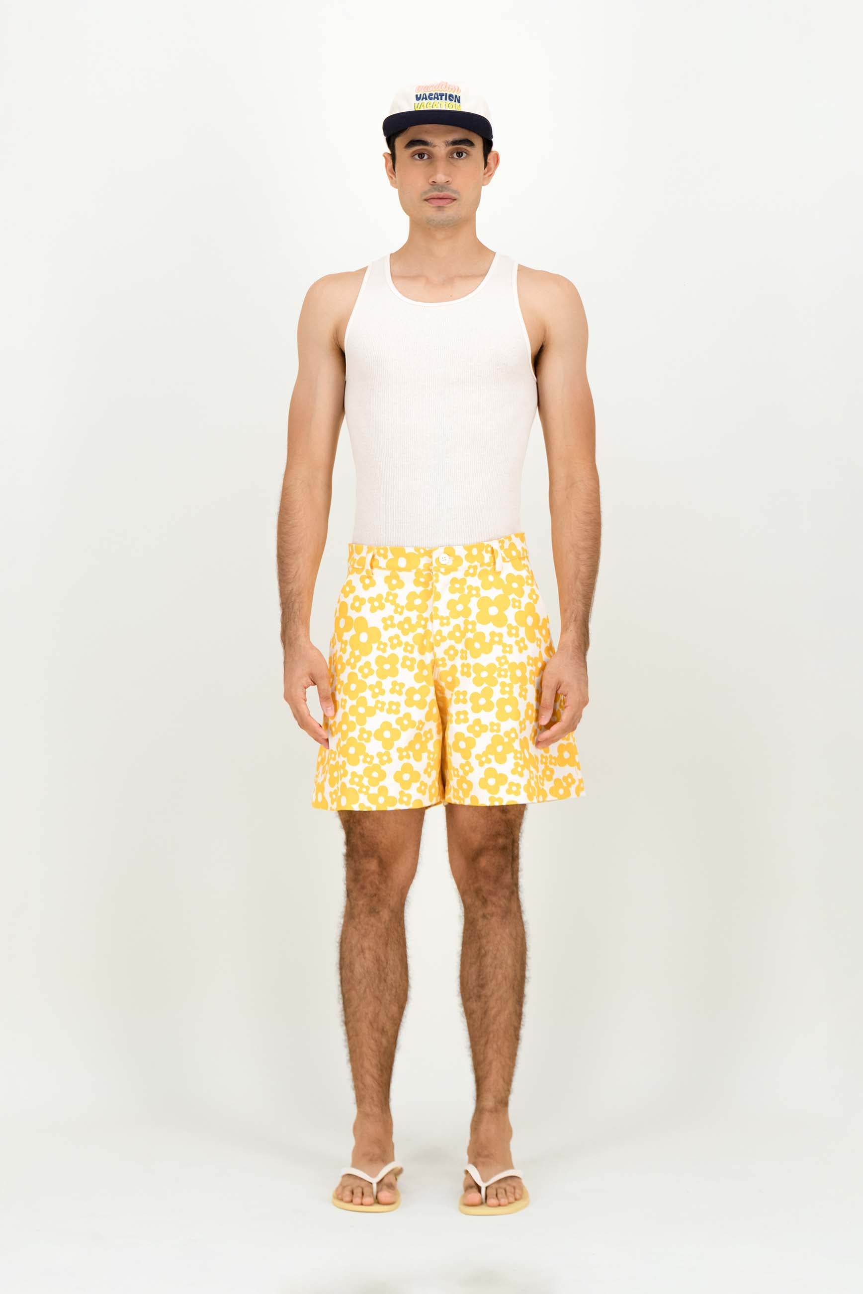 A man modeling a white tank top, yellow and white patterned swim trunks, and a black and white baseball cap against a plain white studio background.