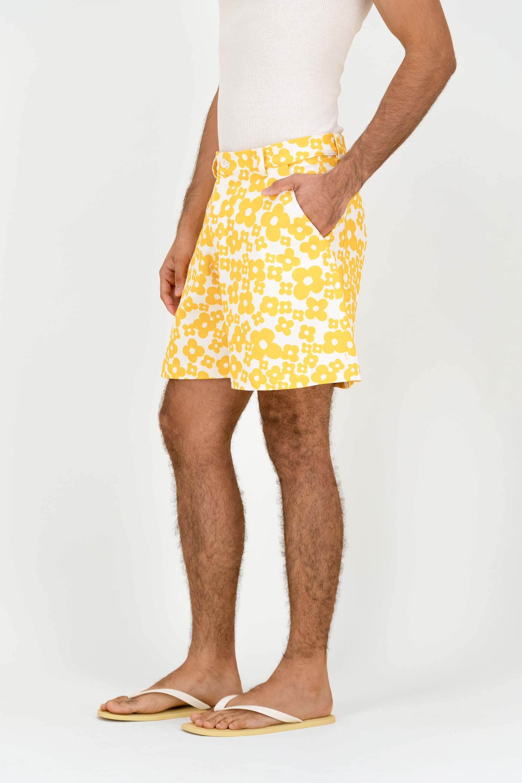 A man wearing yellow and white patterned shorts, a white tank top, and beige flip-flops, standing against a plain white studio background.
