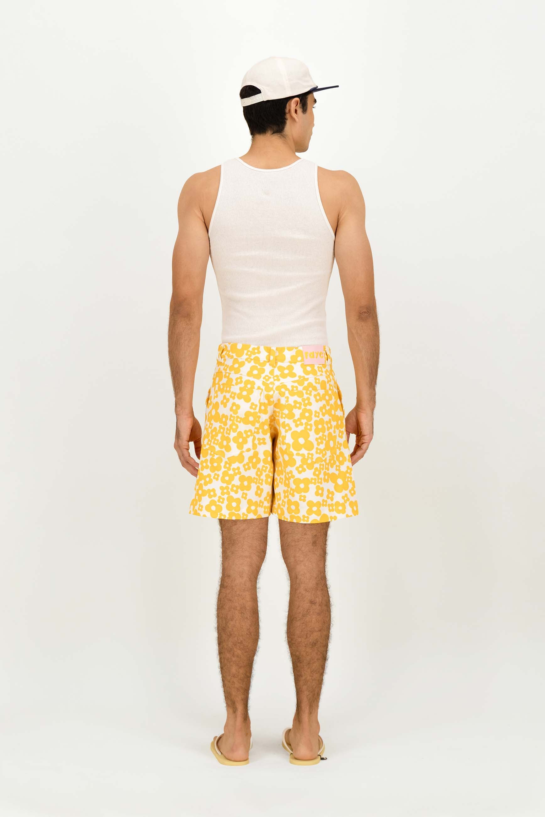 A man is shown from behind wearing yellow and white patterned swim trunks, a white tank top, and a white baseball cap against a plain white studio background.