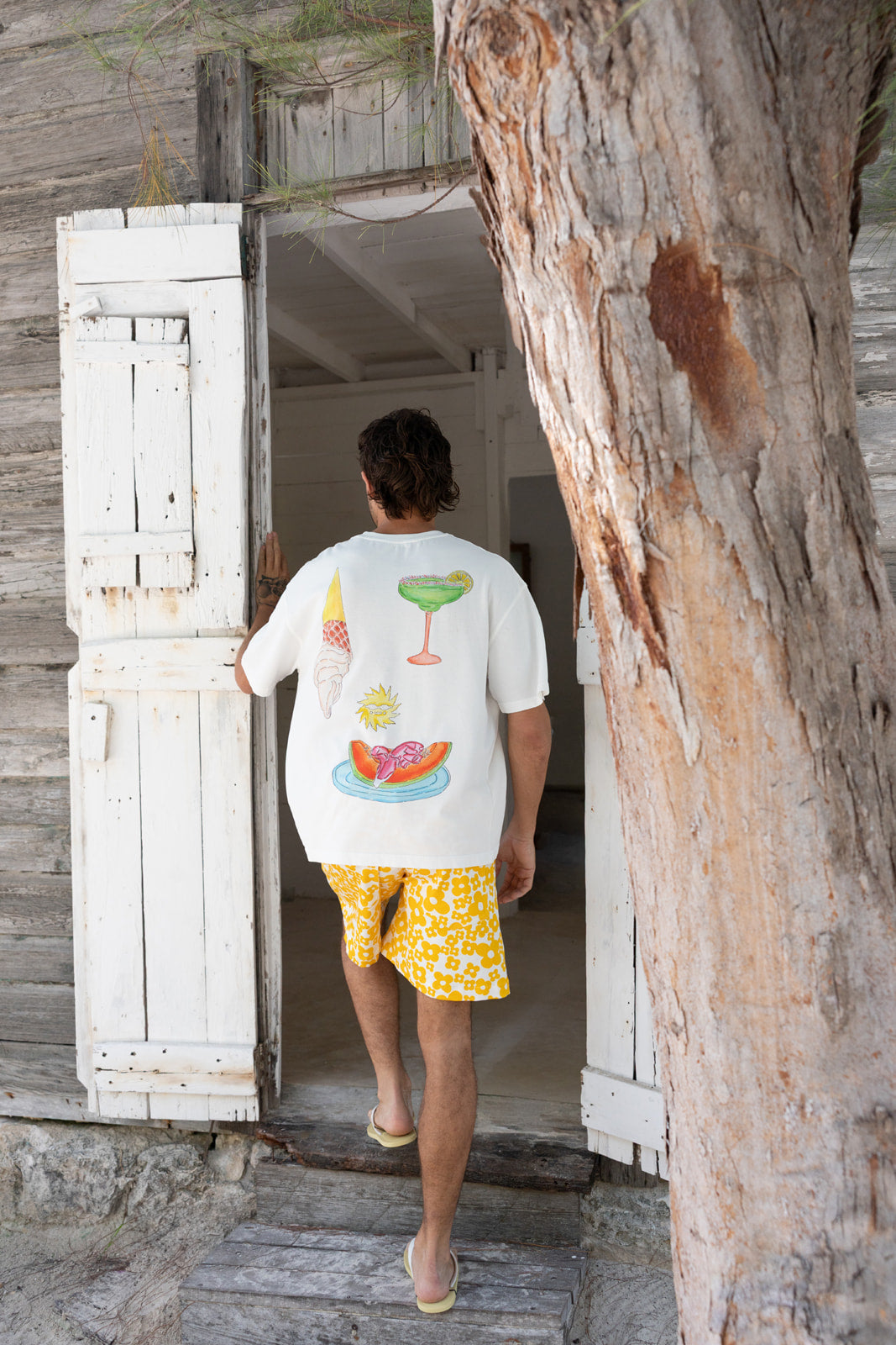 A man wearing a white graphic t-shirt and yellow patterned shorts walks out of a rustic wooden doorway, with a large tree trunk on the right side of the frame.