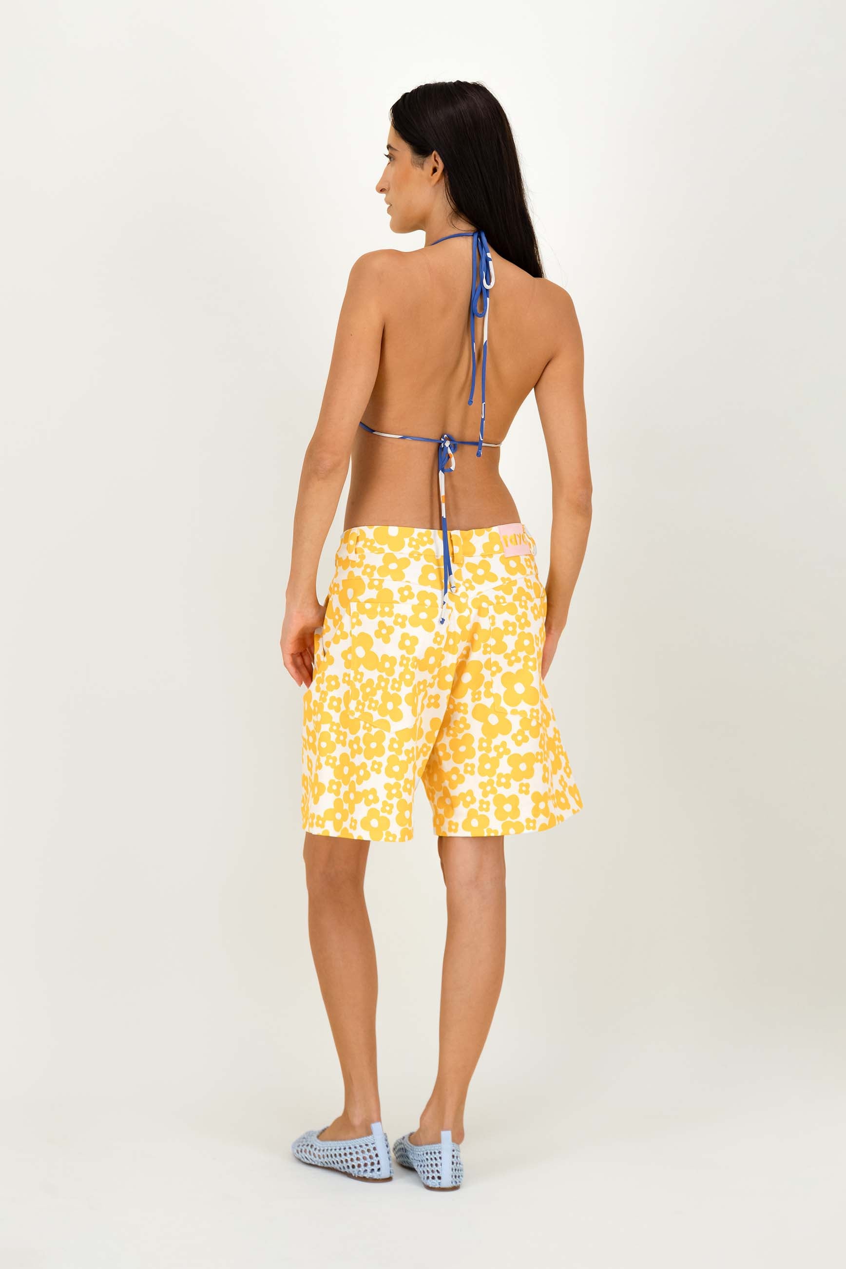 A woman shown from the back wearing yellow and white patterned shorts and a blue string halter top, standing against a plain light gray studio background.