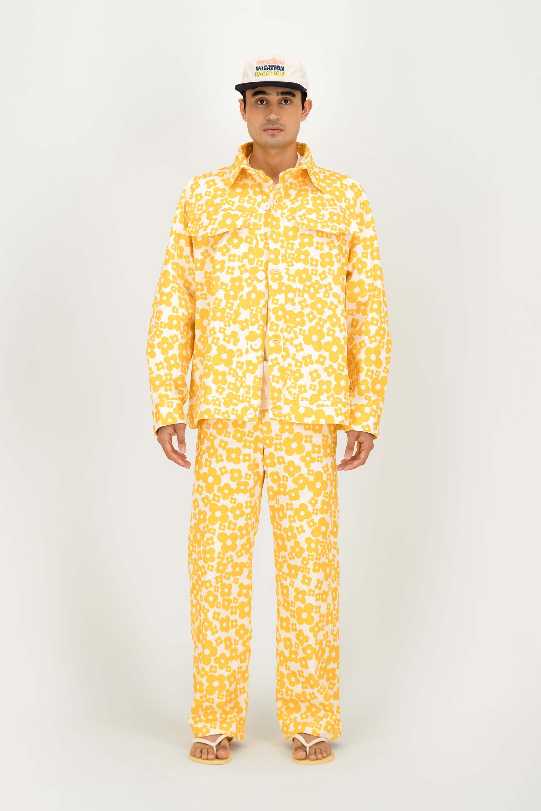 A man modeling a matching two-piece set in a bright yellow and white flower print pattern, paired with a white baseball cap and sandals, against a plain light gray background.