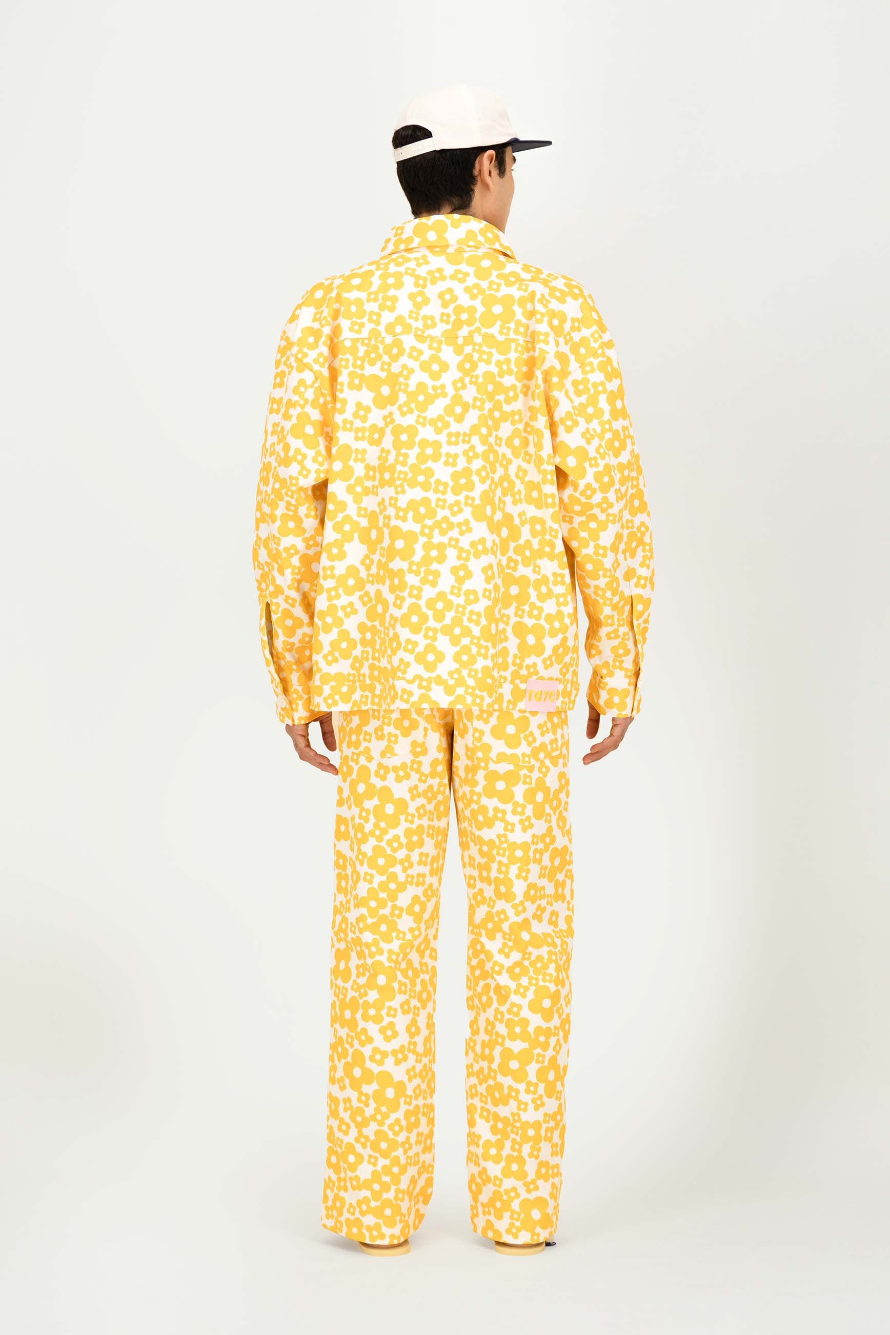 A model viewed from the back wearing a matching yellow and white patterned jacket and trousers set, paired with a white cap, against a plain white studio background.