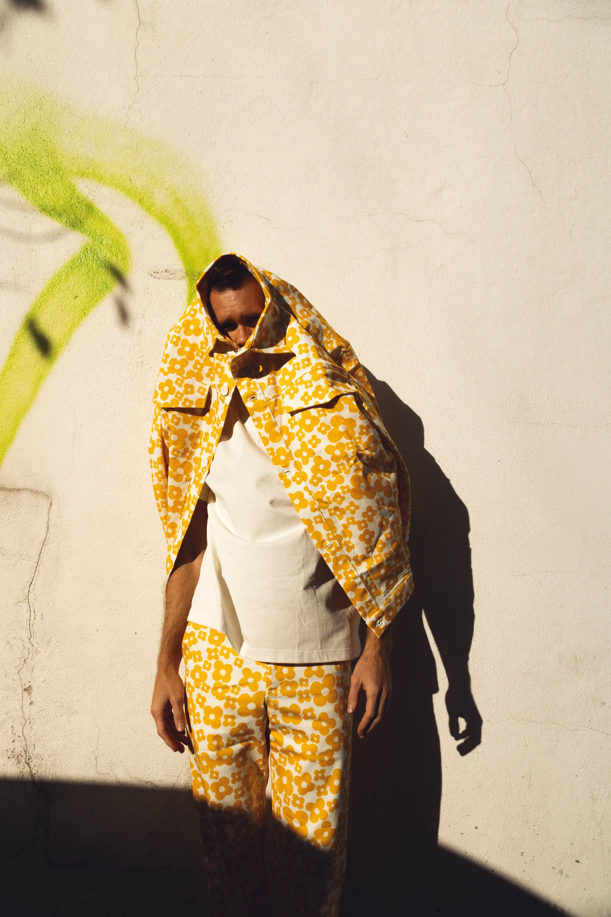 A model wearing a matching yellow and white floral print jacket and trousers, styled over a white t-shirt, standing against a textured off-white wall with a strong shadow.