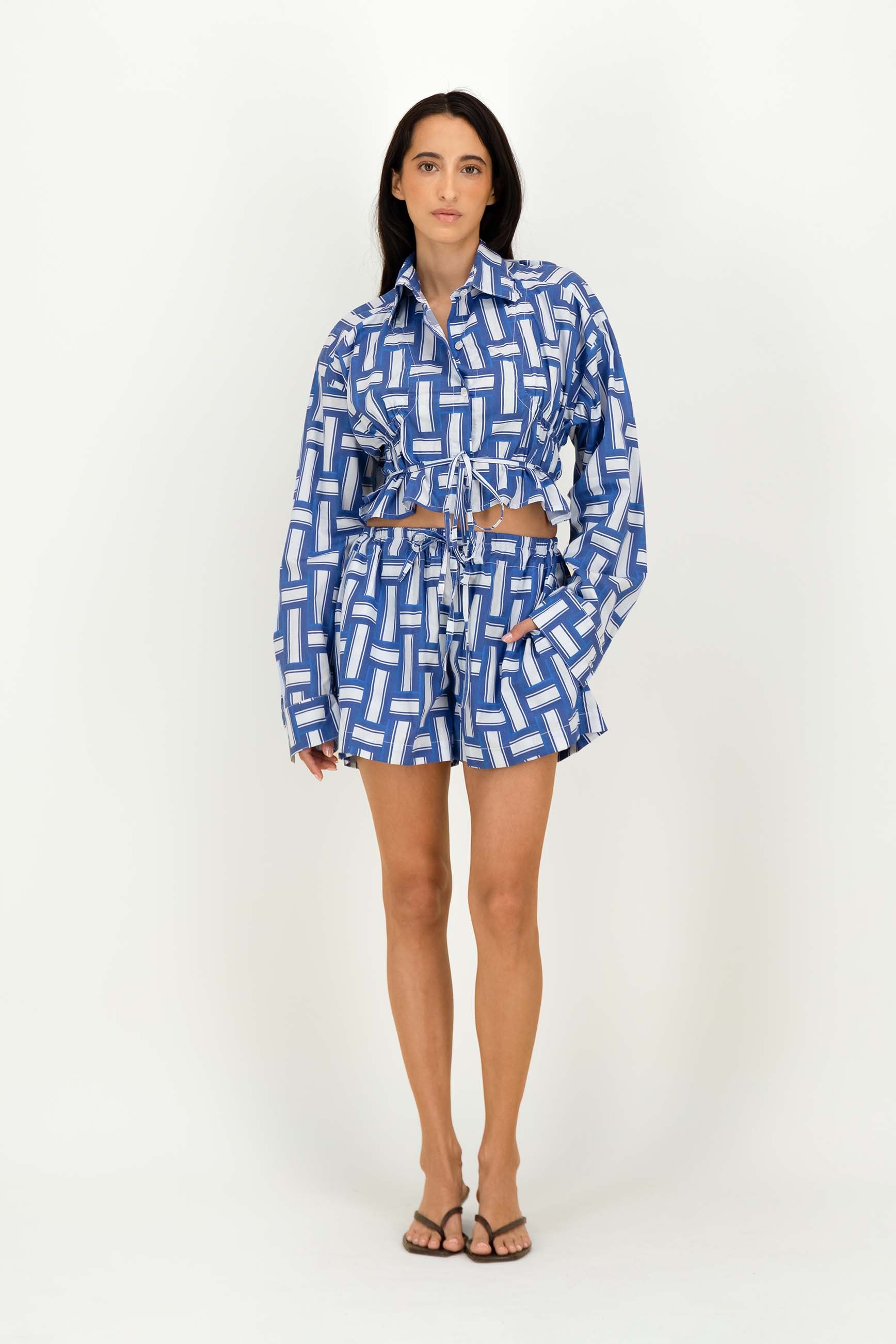 A woman modeling a matching blue and white geometric print two-piece set consisting of a long-sleeve cropped tie-front shirt and matching shorts, paired with brown strappy sandals against a plain white studio background.