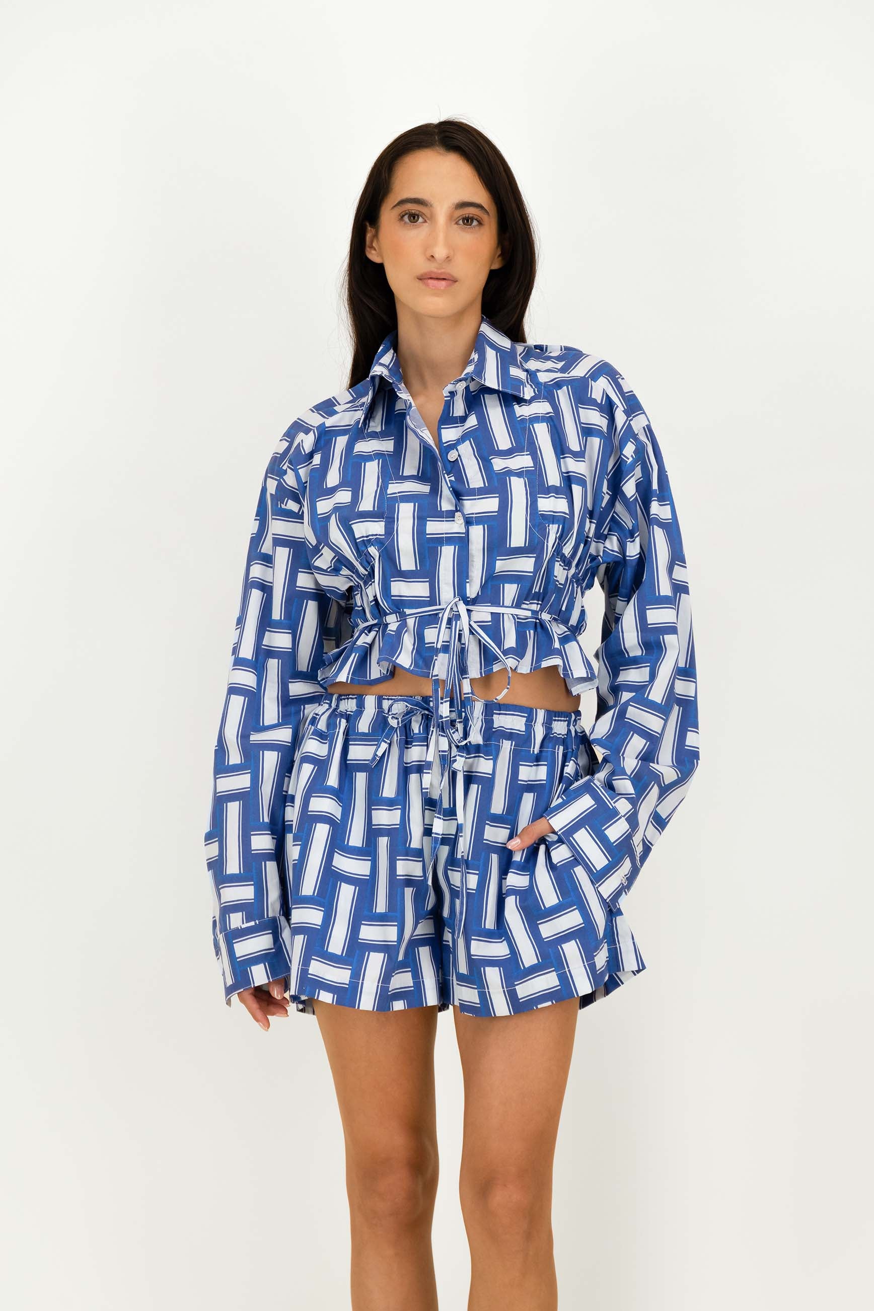 A woman modeling a matching blue and white geometric print long-sleeve crop top and shorts set against a plain white studio background.