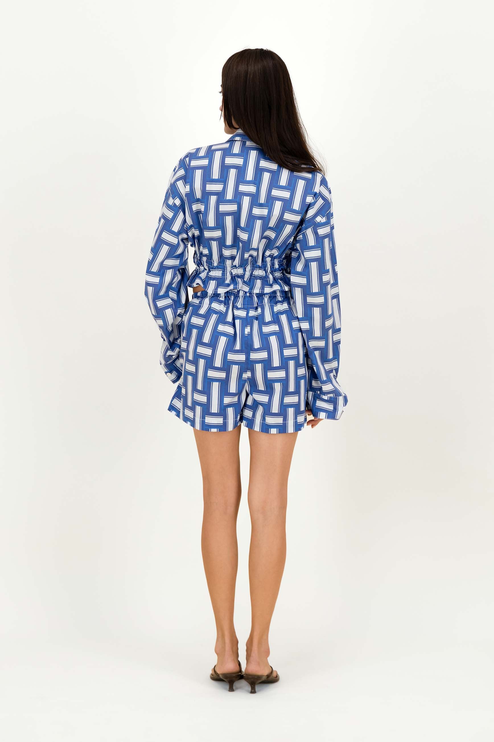 A woman viewed from the back wearing a blue and white geometric patterned romper with long sleeves and a drawstring waist, standing against a plain white studio background.