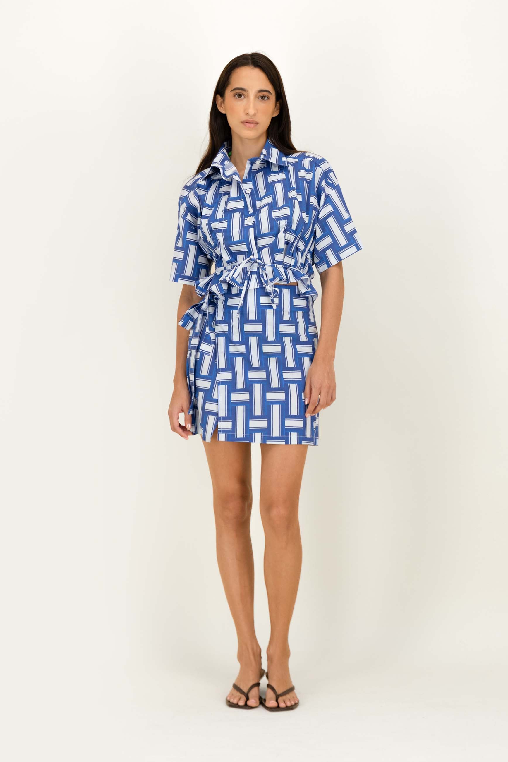 A woman modeling a short-sleeve collared shirt  with a blue and white geometric pattern and a matching tie waist, photographed against a plain light background.