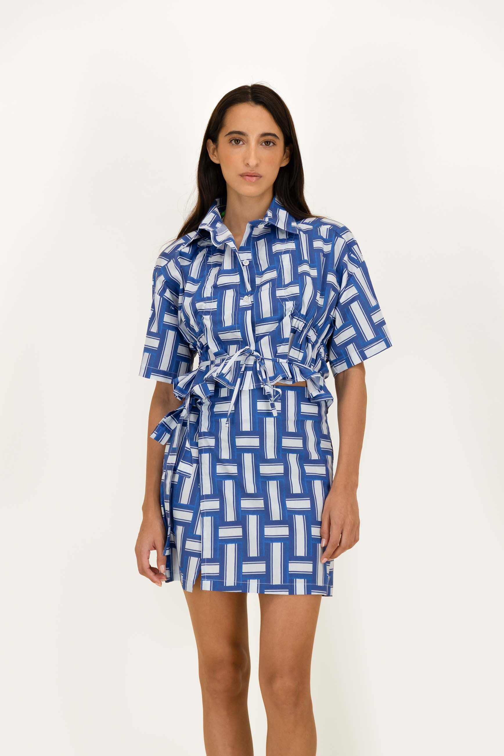 A woman modeling a short-sleeve collared shirt with a blue and white geometric pattern and a matching tie belt, photographed against a plain light background.