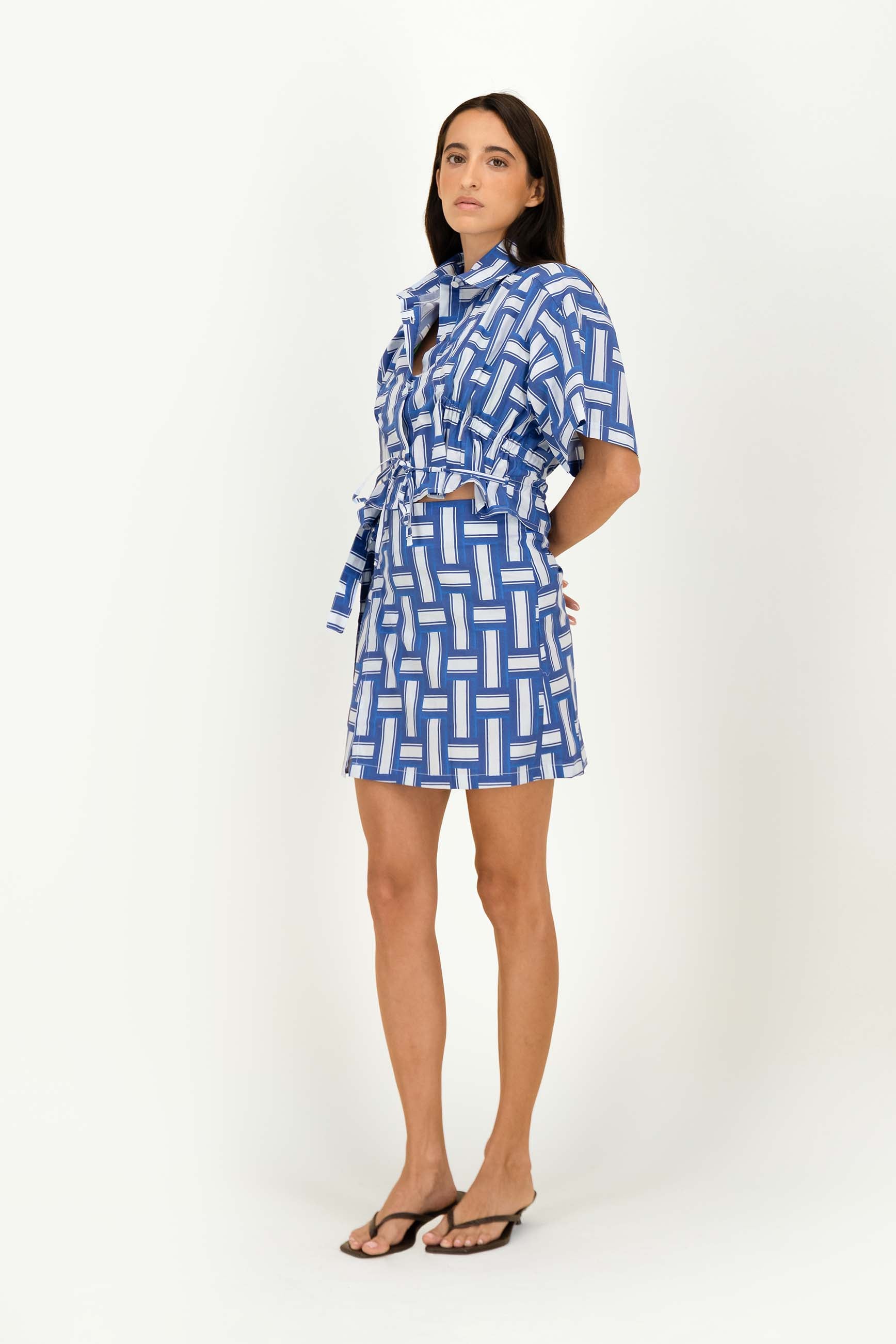 A woman modeling a blue and white geometric patterned shirt with short sleeves, a tie-neck detail, and a matching tie belt, standing against a plain white studio background.