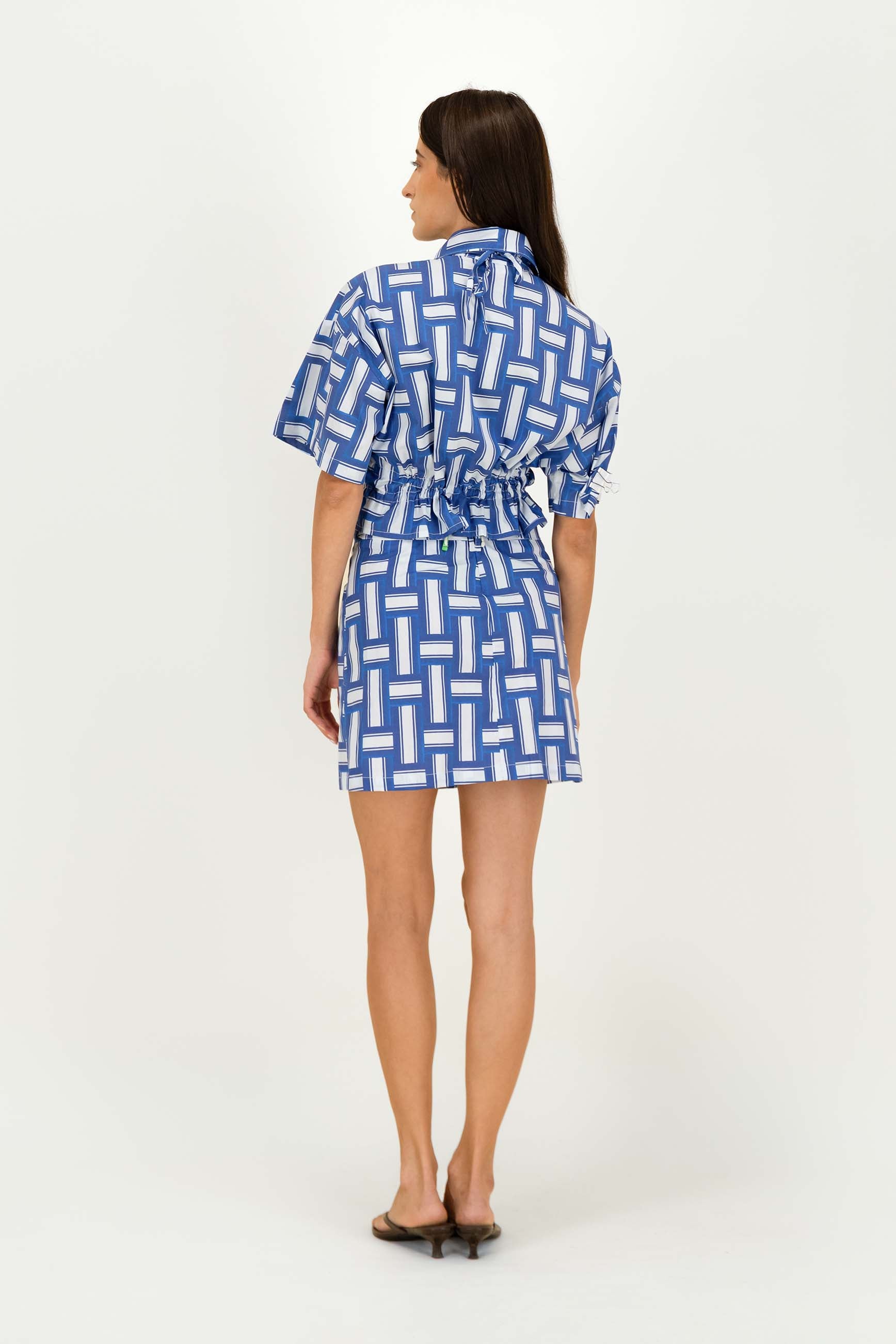 A woman is shown from the back wearing a short-sleeved shirt with a blue and white geometric pattern, photographed against a plain white studio background.