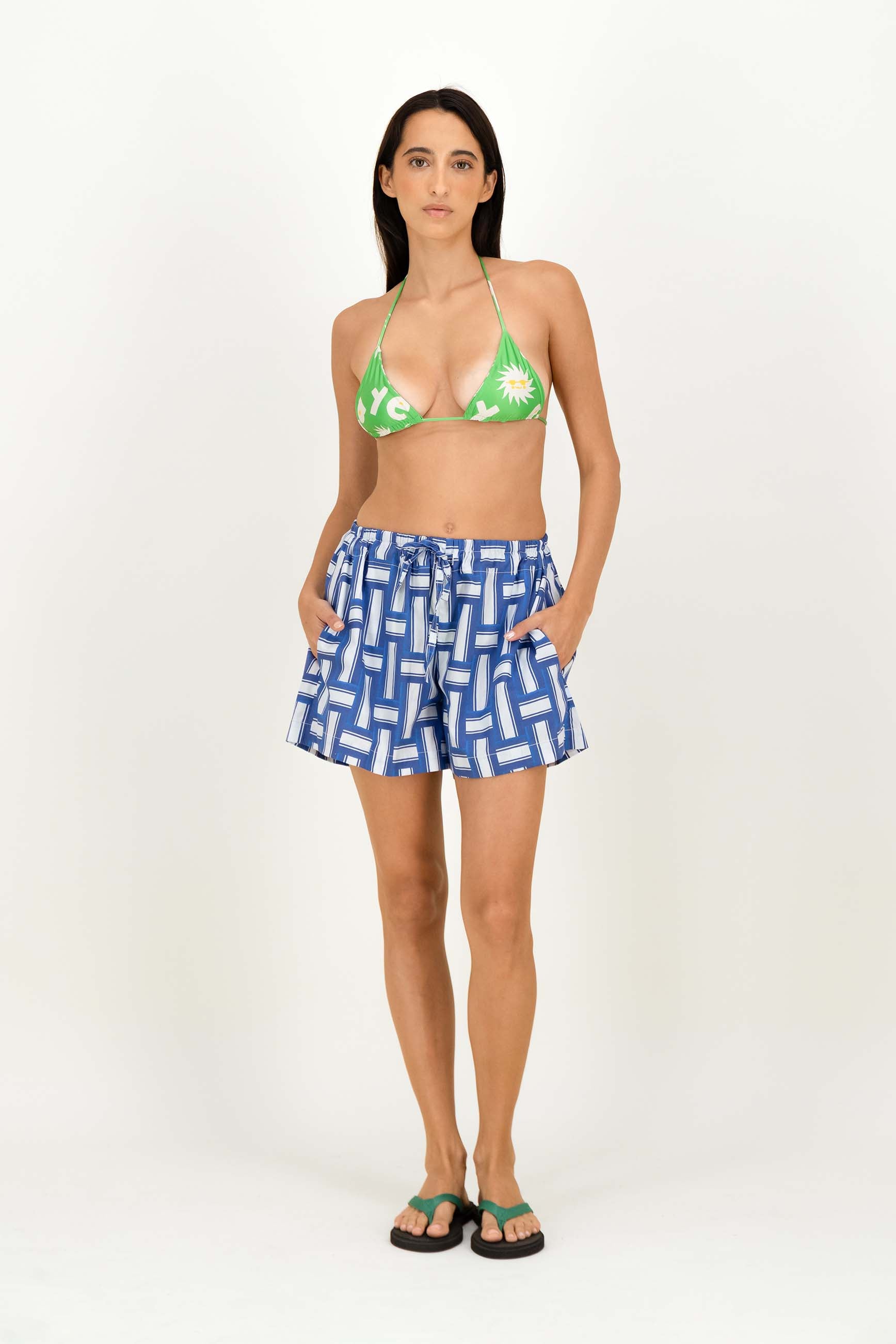 A woman modeling a swimwear set consisting of a green triangle bikini top with white floral patterns and blue-and-white geometric patterned shorts, paired with green flip-flops, against a plain white studio background.