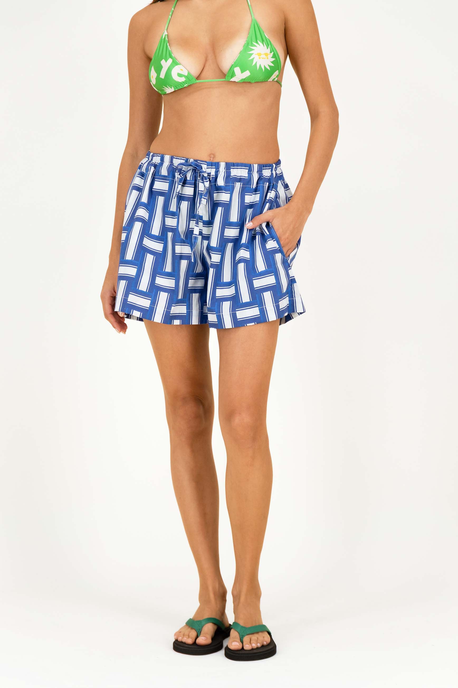 A model wearing blue and white geometric patterned swim shorts with a drawstring waist, paired with a green bikini top and green flip-flop sandals, against a plain white studio background.