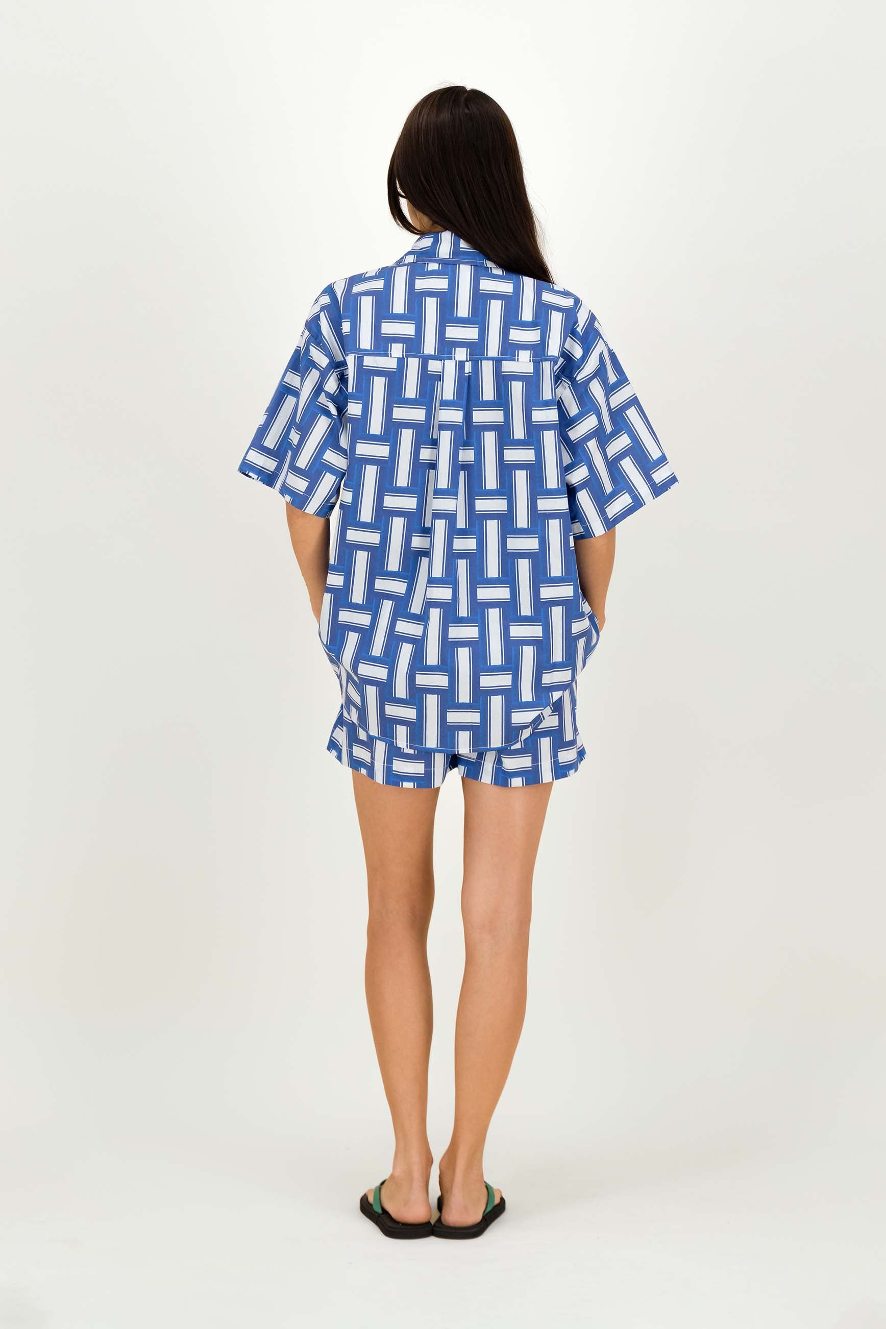 A woman viewed from behind wearing a blue and white geometric patterned short-sleeve shirt and matching shorts, standing against a plain light background.