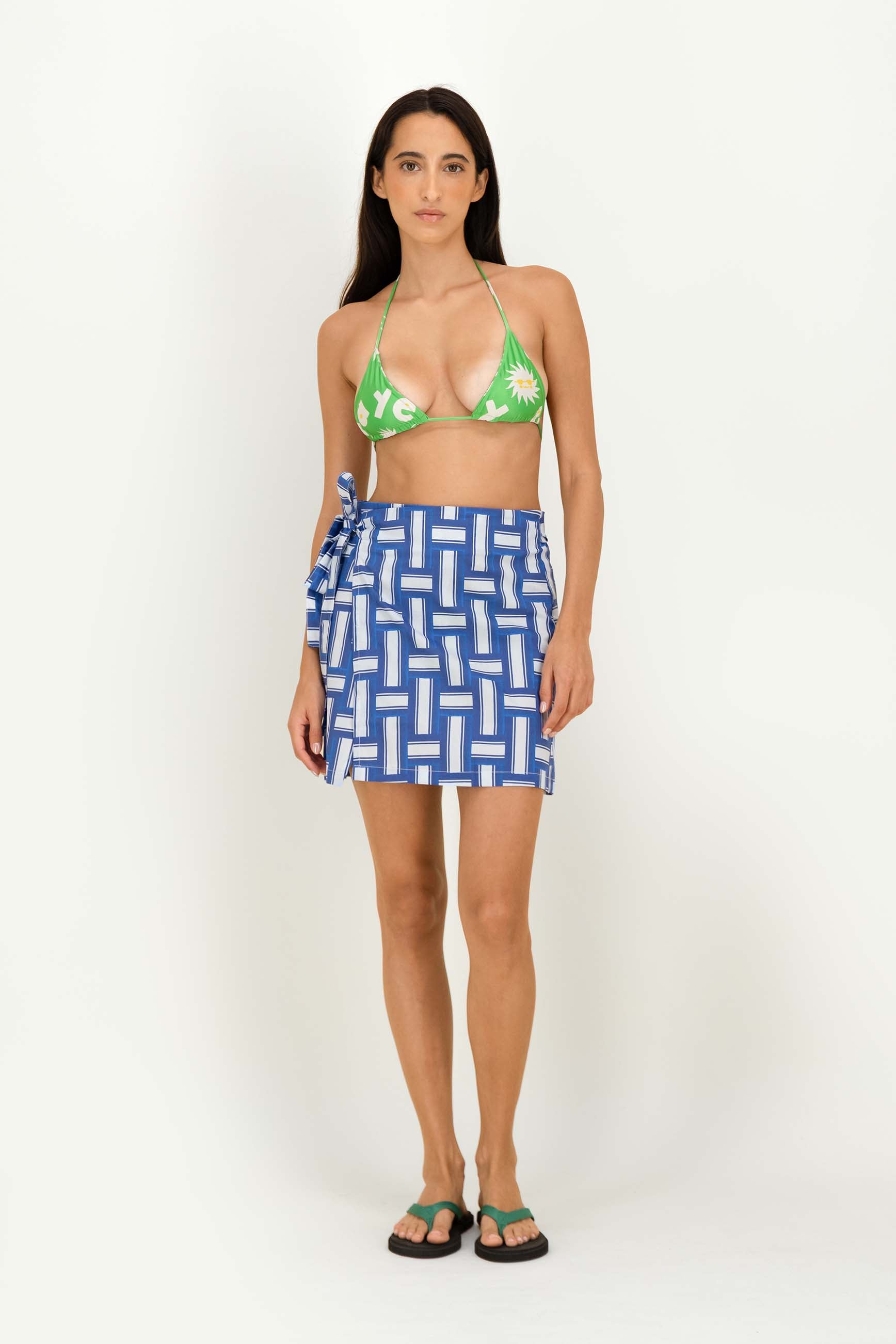 A woman modeling a green floral triangle bikini top paired with a blue and white geometric patterned wrap wrapping skirt, standing against a plain white studio background.