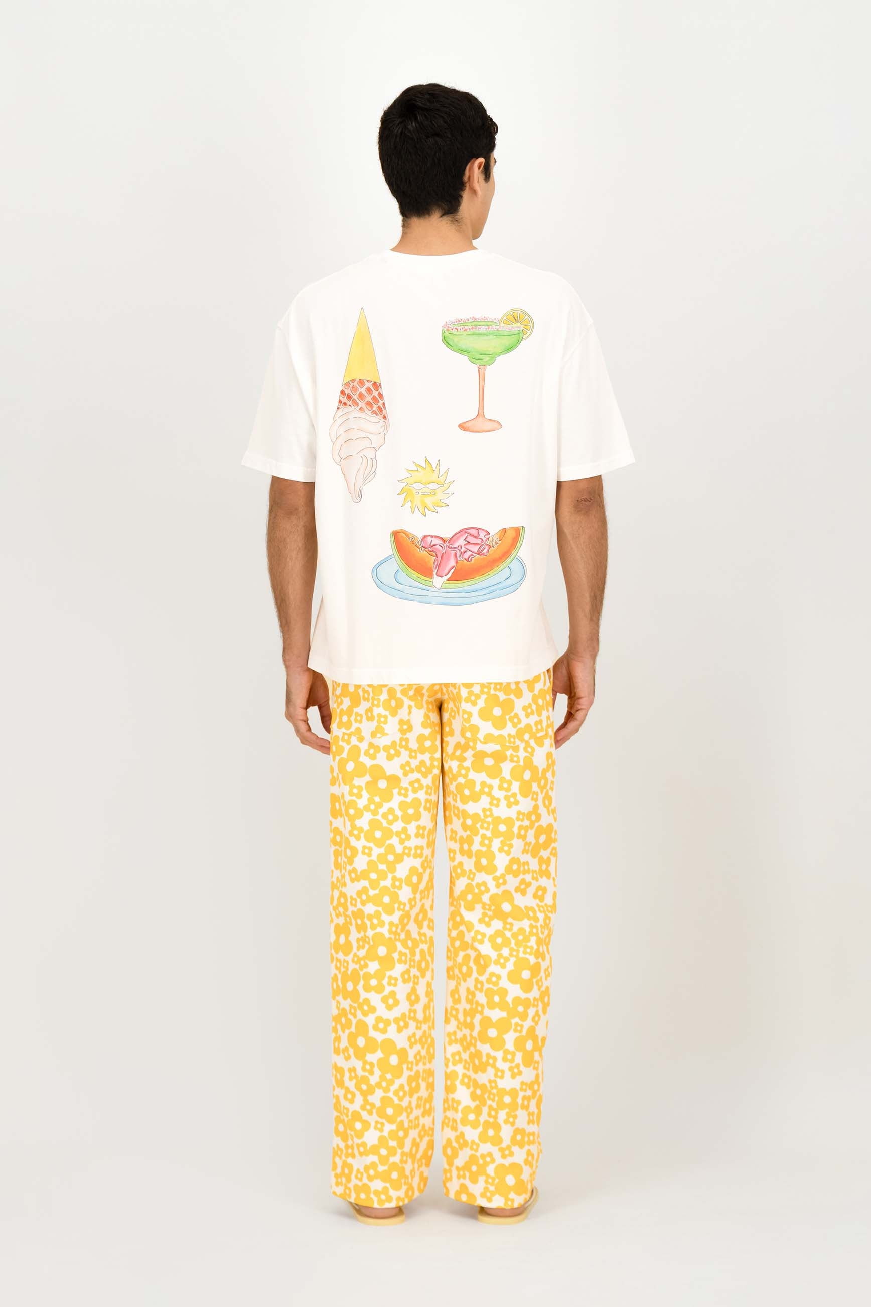 A man viewed from the back wearing a white graphic t-shirt with colorful food illustrations and yellow patterned pants, set against a plain light gray studio background.