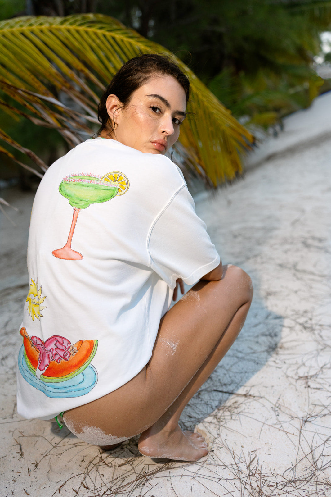 A woman crouching on a sandy beach wearing a white oversized t-shirt with colorful tropical cocktail and watermelon graphics on the back.