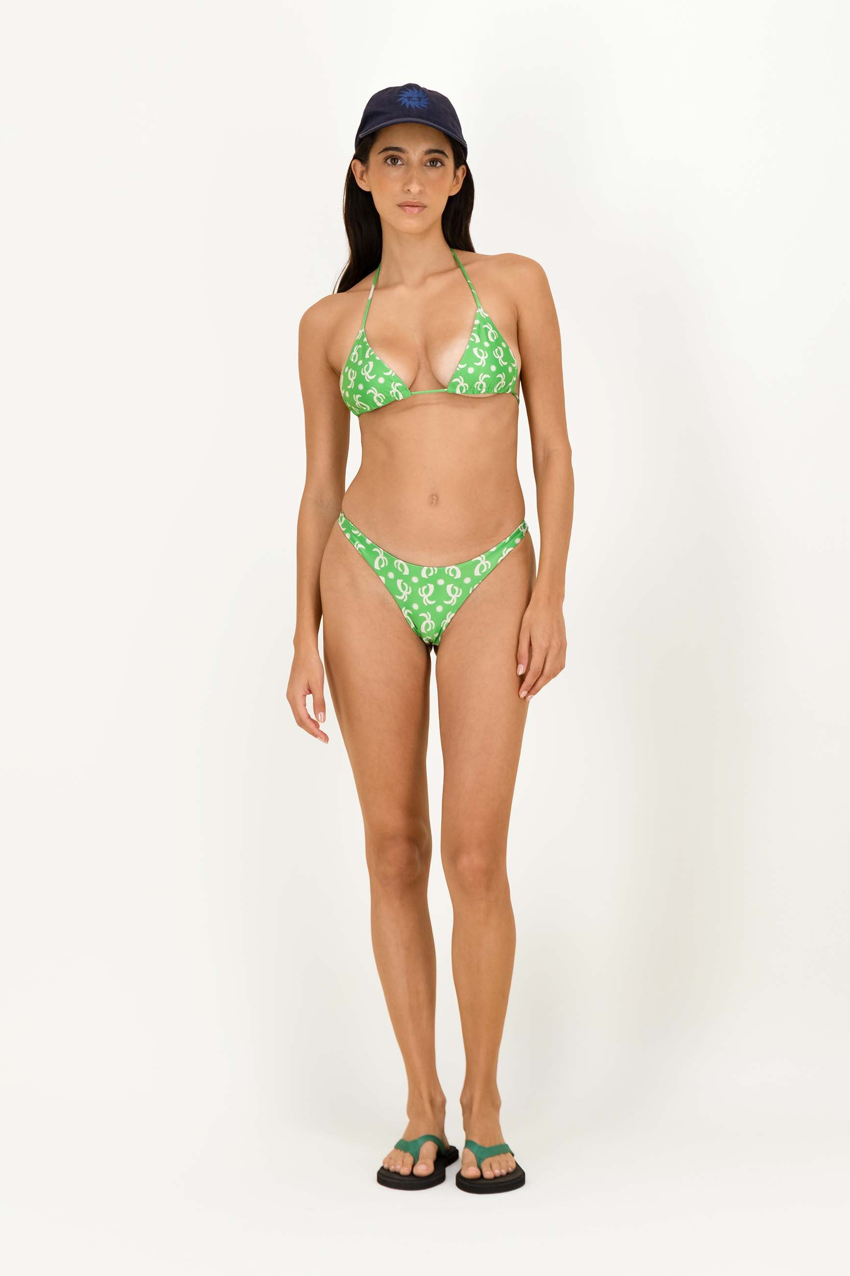 A woman modeling a green and white patterned two-piece bikini with a dark blue baseball cap and green flip-flops against a plain white studio background.