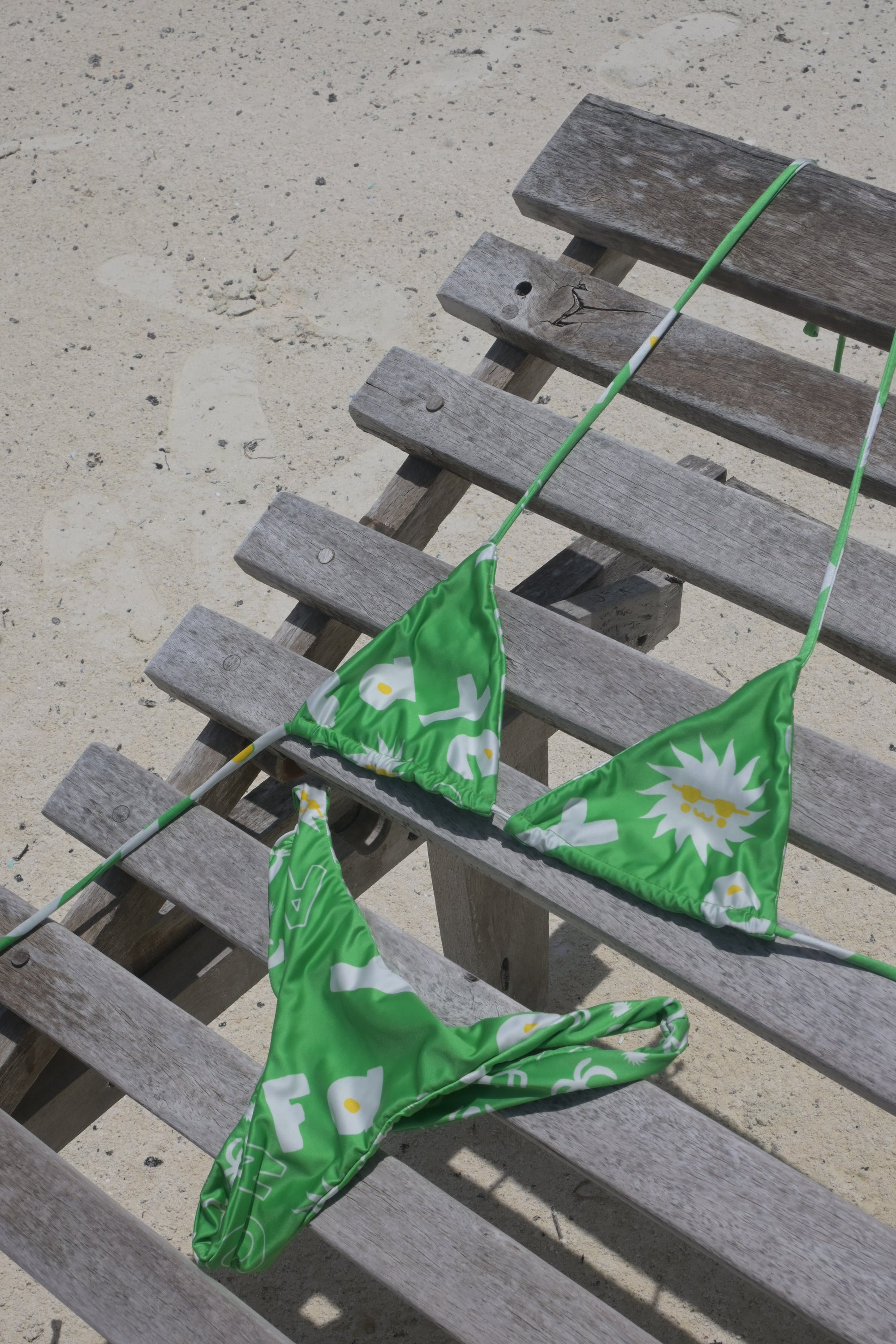 Green string bikini top and bottom with a white daisy print, draped over a weathered wooden beach chair on sand.