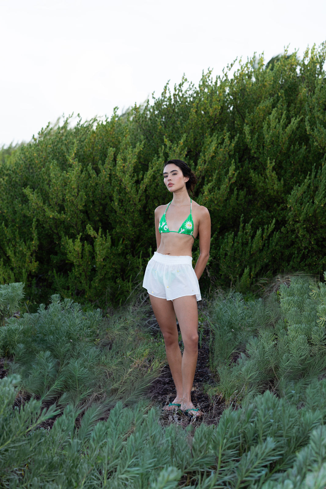 A woman stands outdoors in a field of green plants, wearing a green and white patterned triangle bikini top and white shorts.