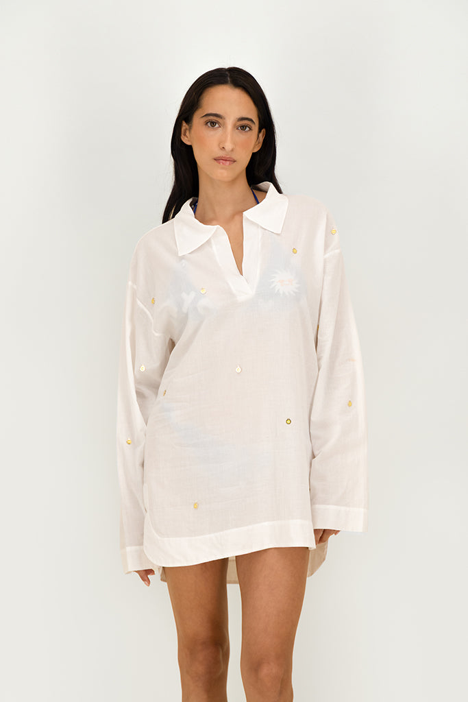 A woman modeling a white long-sleeve collared shirt dress with subtle tonal floral embroidery and small gold medaillon accents, set against a plain light gray studio background.