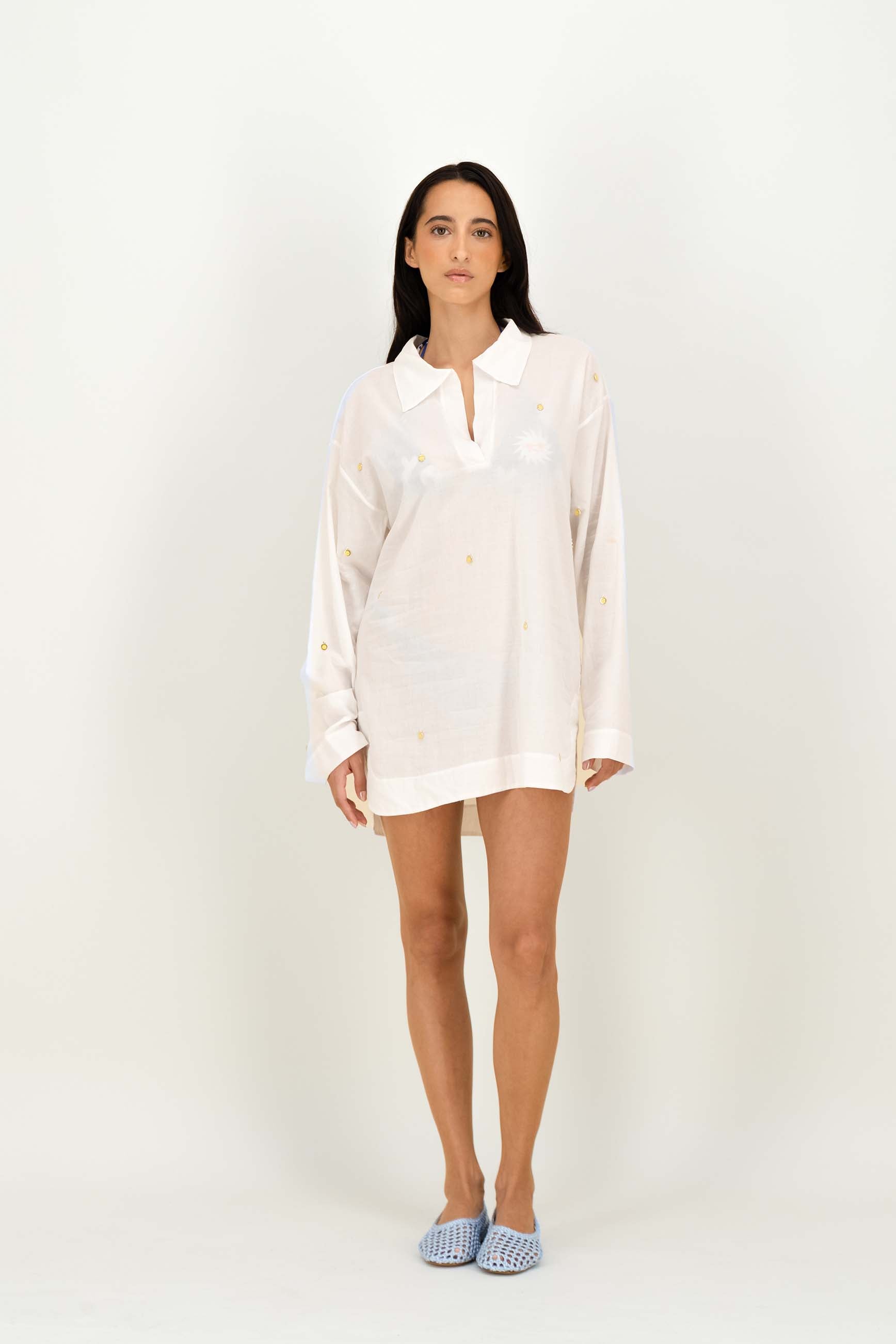 A woman modeling a white long-sleeve tunic with embroidered details, standing against a plain white studio background.
