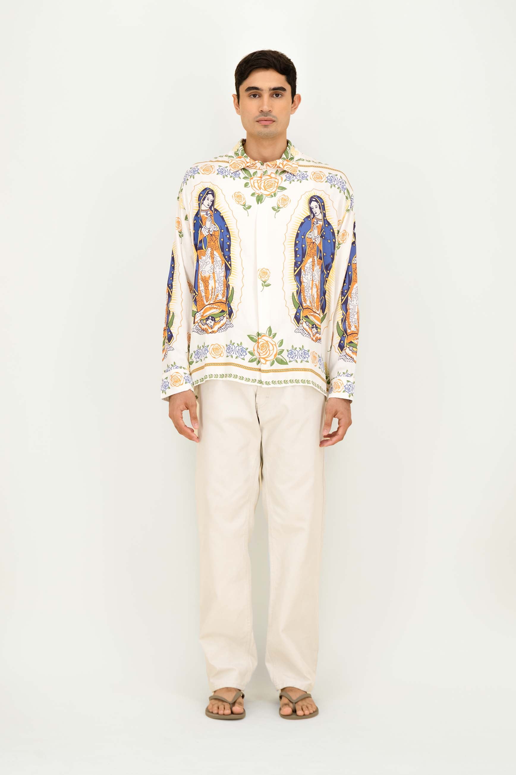 A man modeling a long-sleeve button-up shirt with colorful religious figures and floral patterns on an off-white base, paired with off-white trousers and sandals, against a plain white studio background.