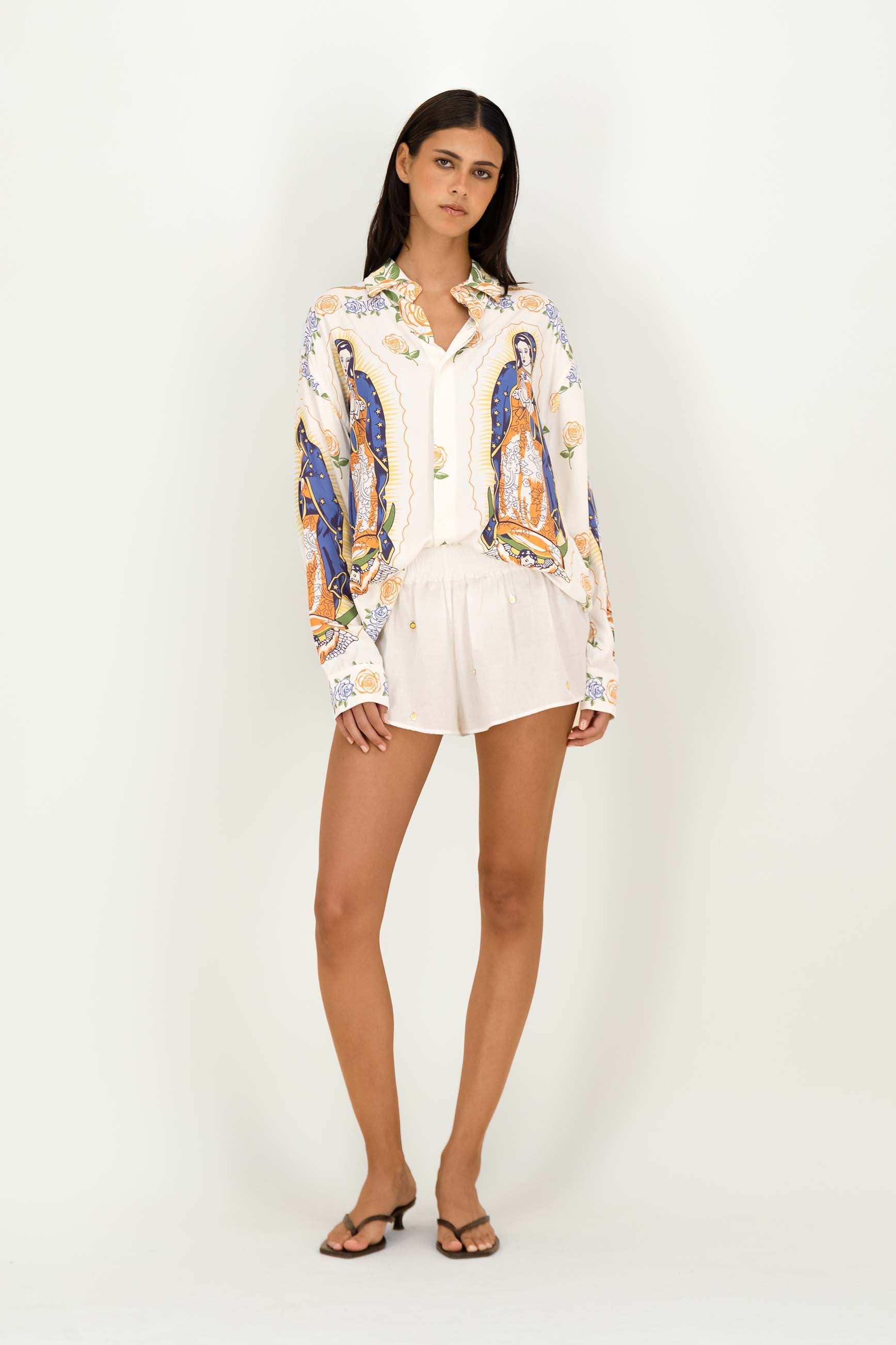 A woman modeling a white button-up shirt with a colorful religious print and white shorts against a plain white studio background.