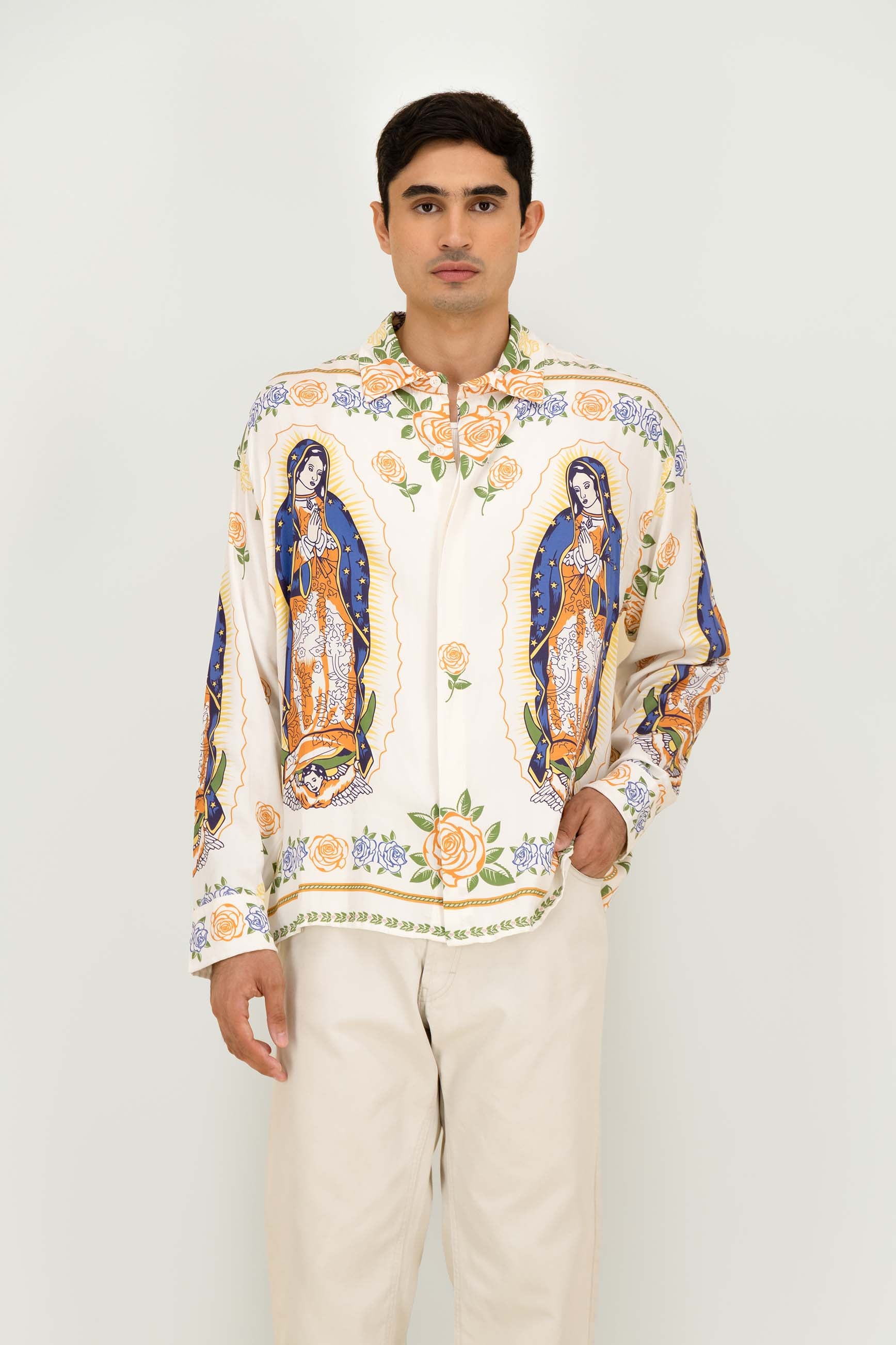 A man wearing a cream-colored long-sleeve button-up shirt with colorful religious prints of Our Lady of Guadalupe and floral motifs, paired with light beige trousers, against a plain light gray background.