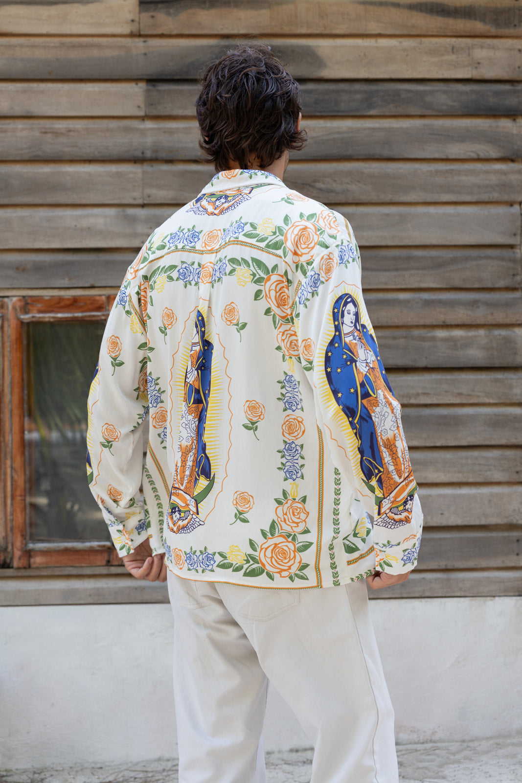 A man viewed from behind wearing a cream-colored long-sleeve shirt with a colorful Mexican folk art print featuring Our Lady of Guadalupe and roses, paired with off-white trousers, standing in front of a rustic wooden wall.