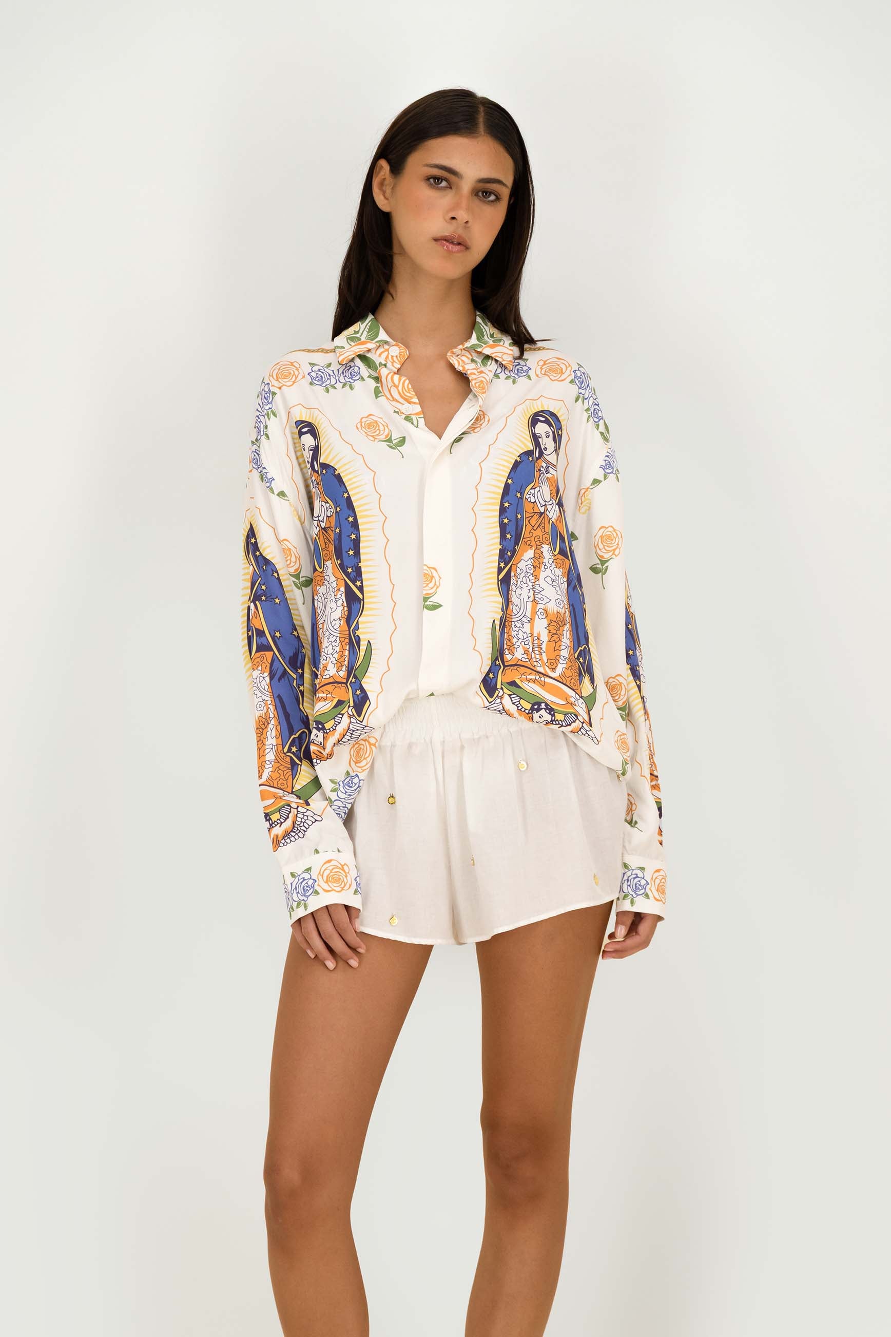 A model wearing a cream-colored button-up shirt with a colorful, detailed religious print and matching cream shorts, standing against a plain light gray background.