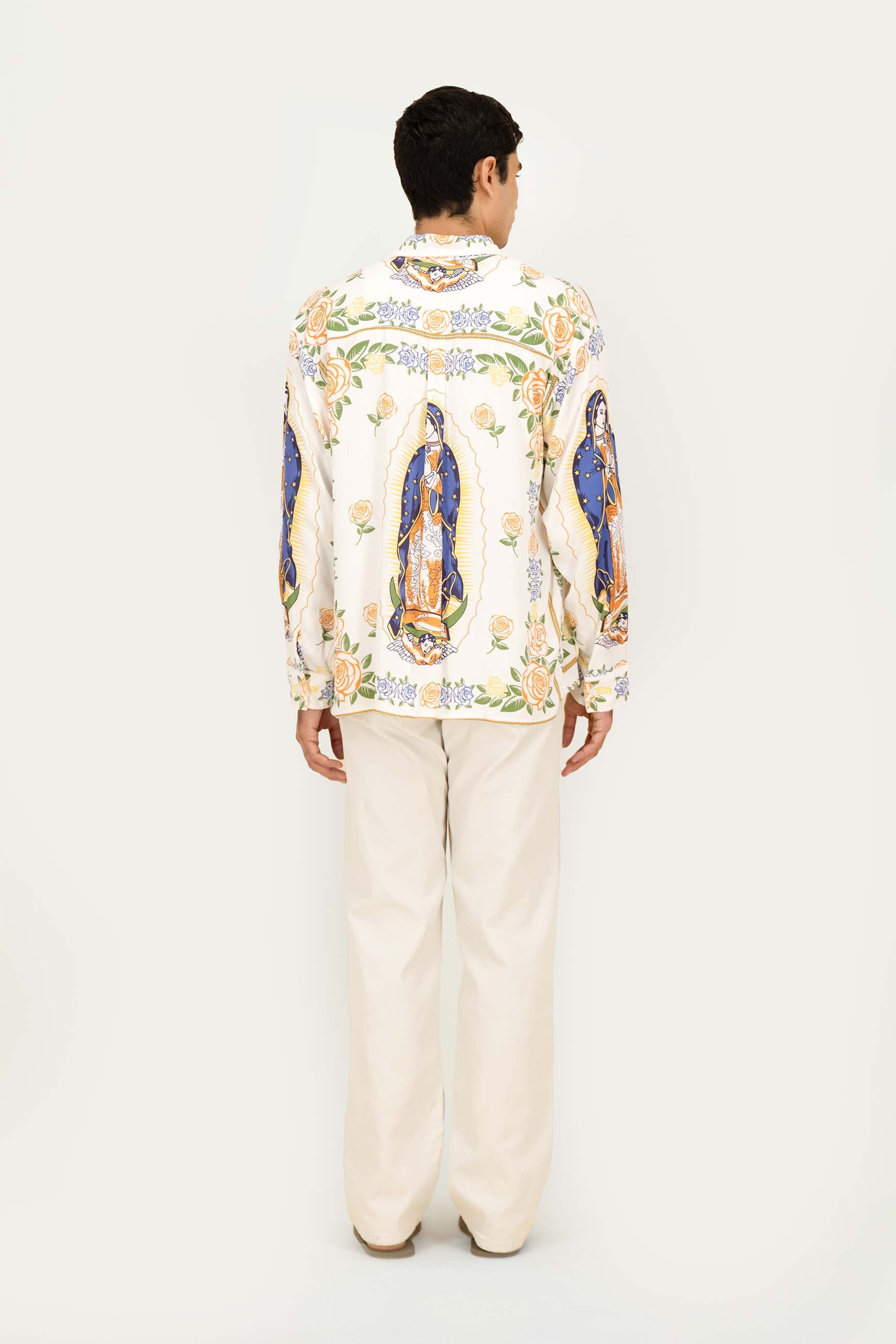 A man viewed from the back wearing a cream-colored long-sleeve shirt with colorful religious prints of Our Lady of Guadalupe, paired with cream-colored trousers, against a plain white studio background.