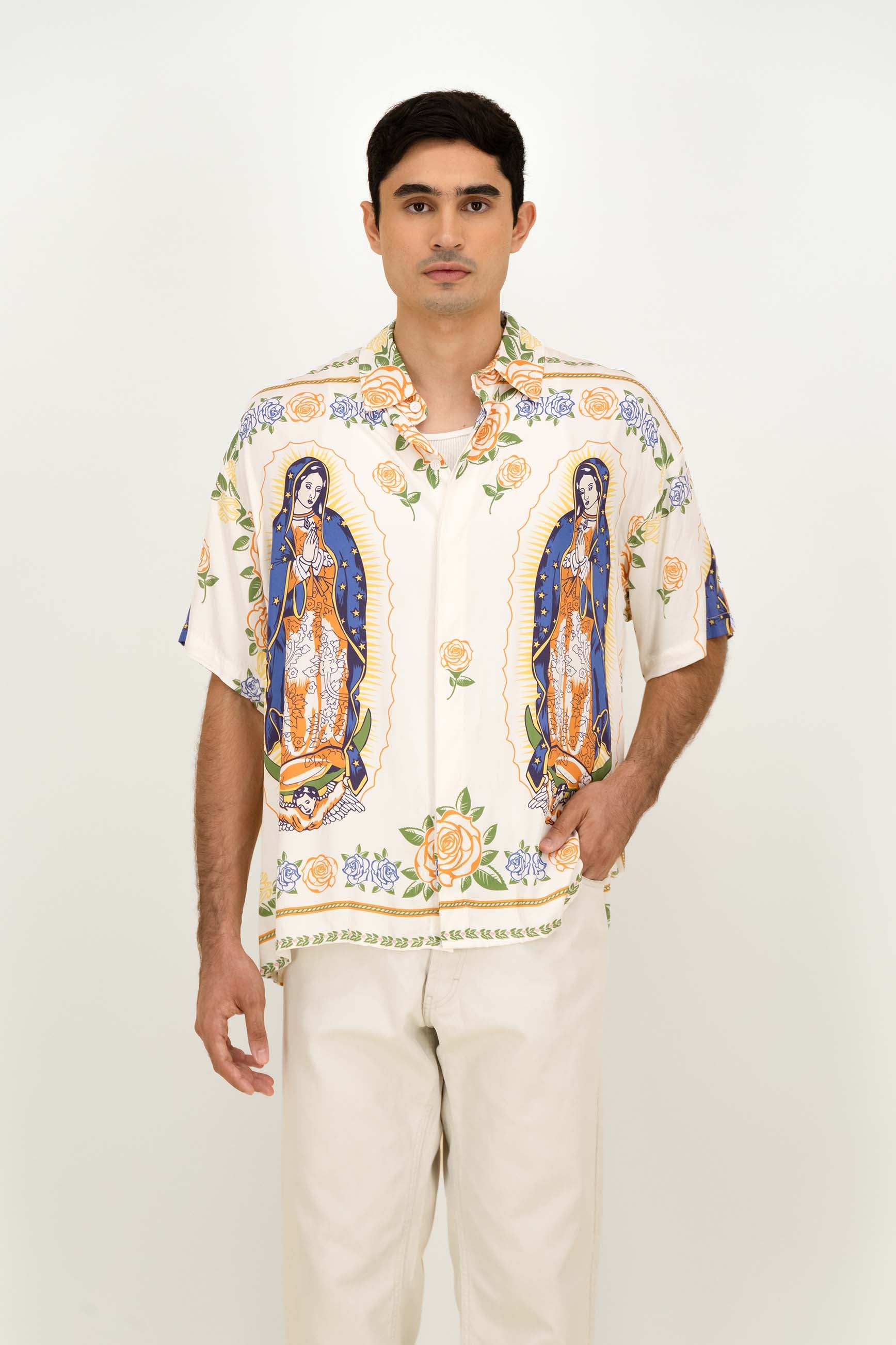 A man modeling a short-sleeve button-up shirt with a white base and colorful religious prints of Our Lady of Guadalupe, paired with light beige trousers, against a plain white studio background.