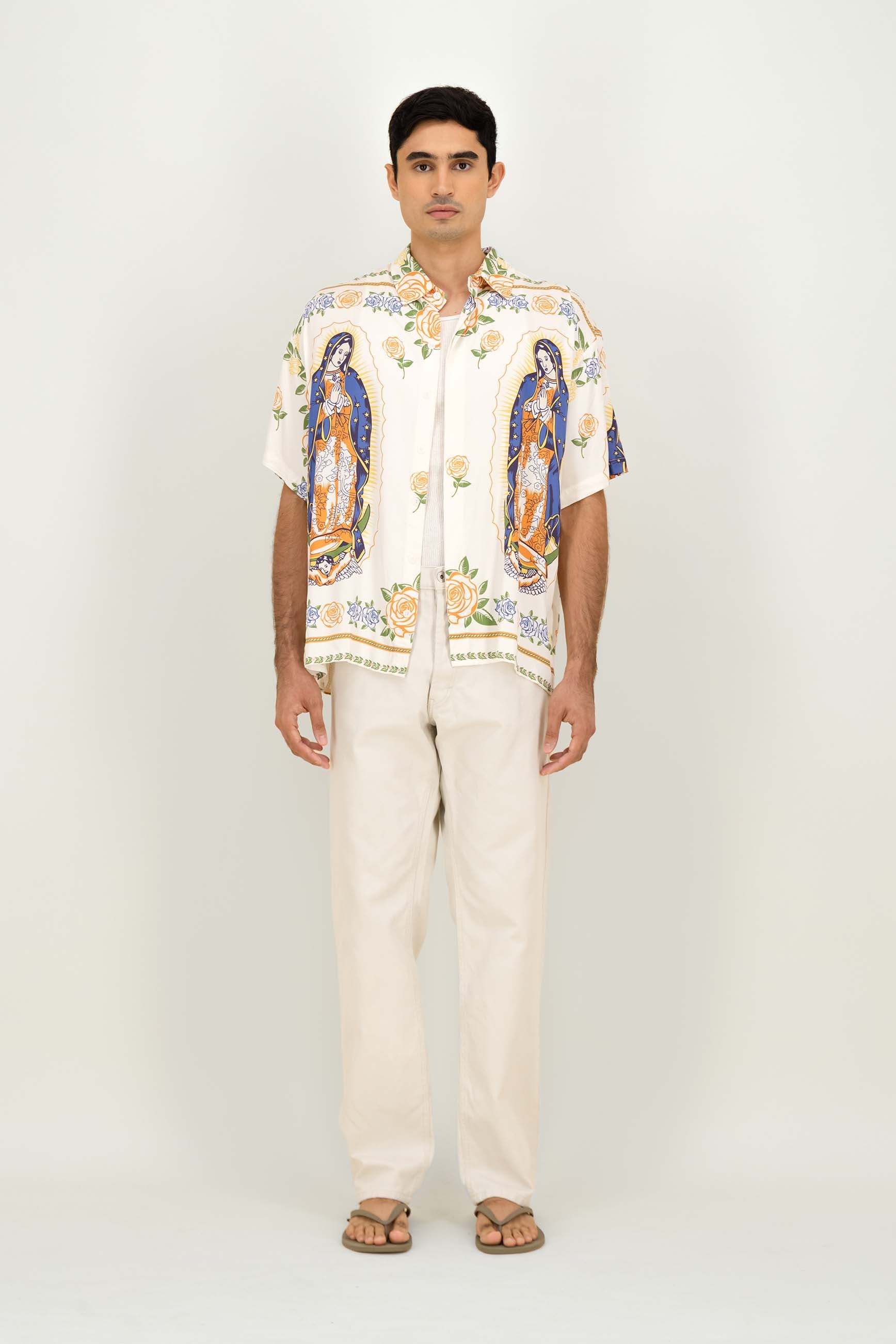 A man modeling a short-sleeve button-up shirt with a colorful religious print of the Virgin Mary, paired with cream pants and sandals, against a plain light gray studio background.