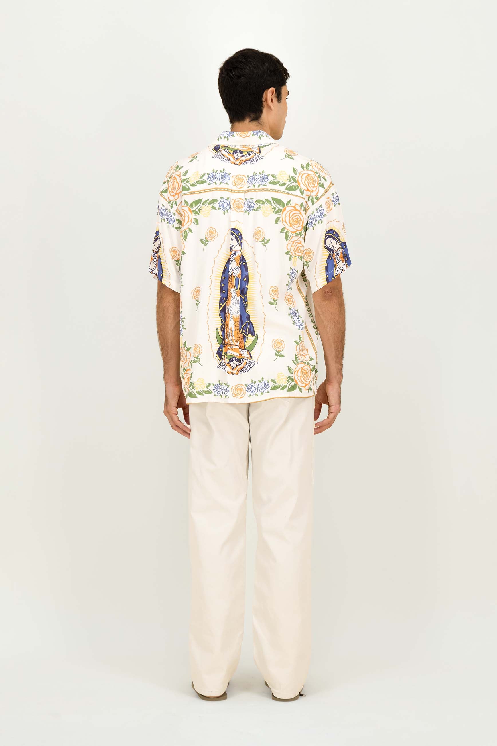A man viewed from behind wearing a cream-colored short-sleeve button-up shirt with a colorful Mexican folk art print of Our Lady of Guadalupe, paired with cream-colored trousers, against a plain white studio background.